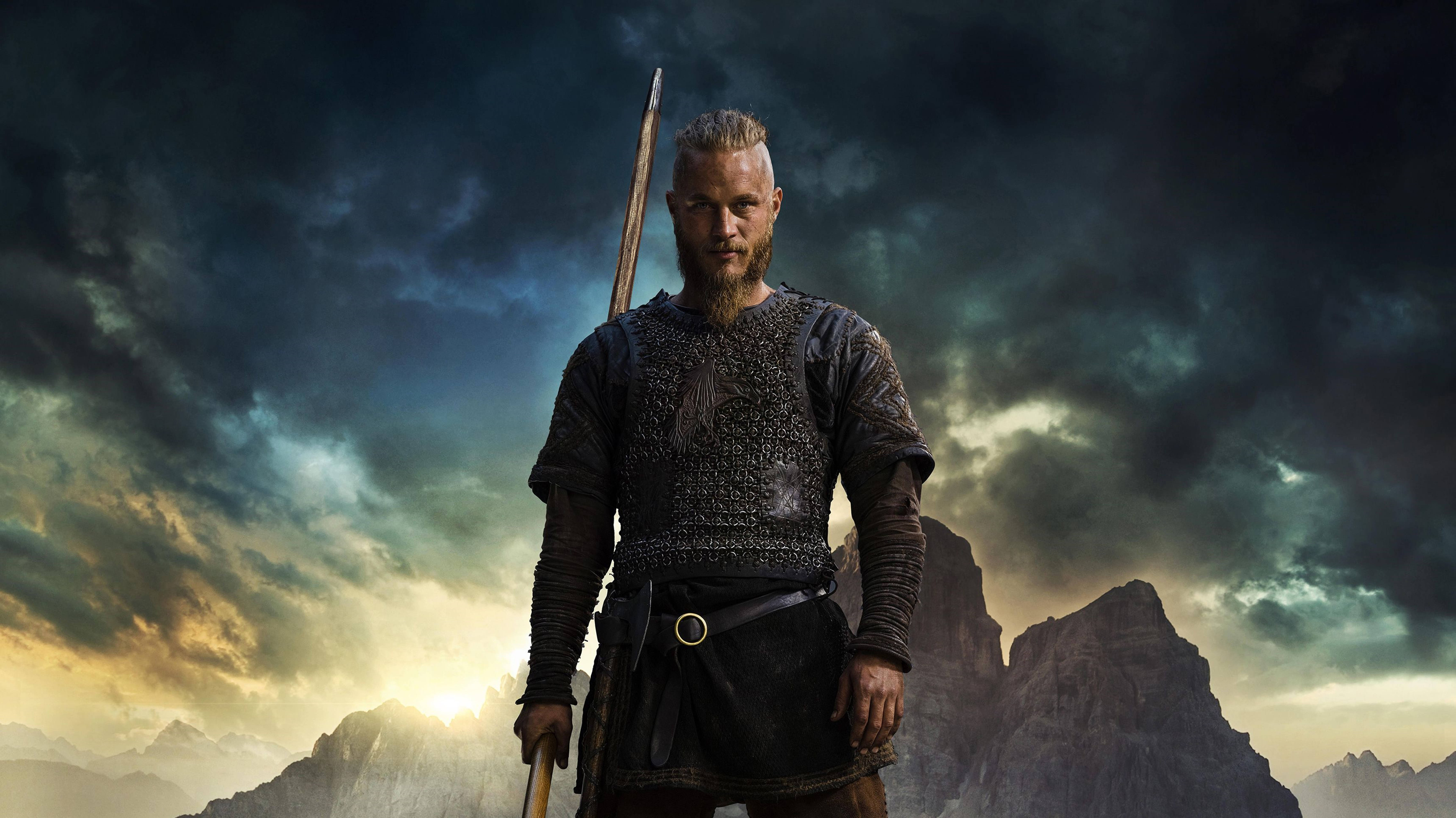 Vikings 桌布 壁紙 — 一位留著鬍鬚、戴著角盔的兇猛維京戰士，在暴風雨的天空下，以戲劇性的山脈為背景，沐浴在金色的日落光芒中。