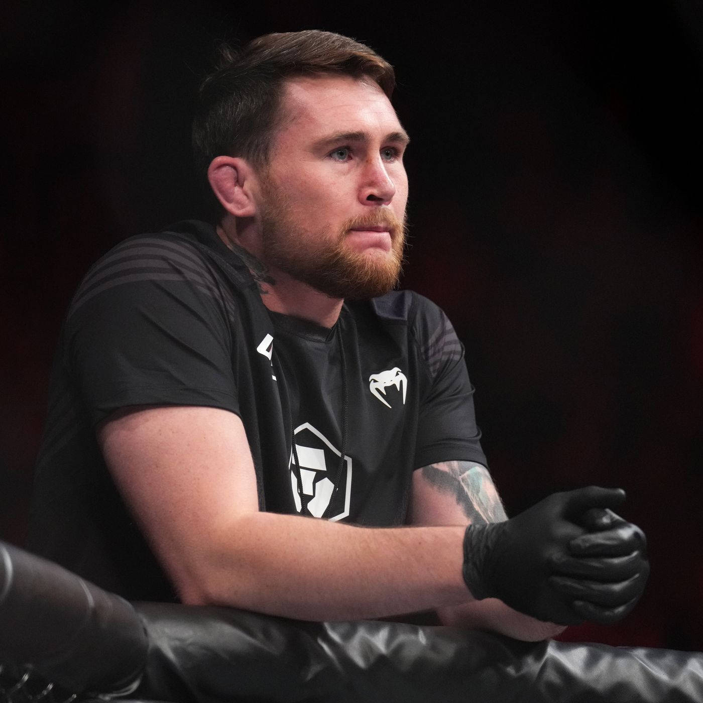 UFC 桌布 壁紙 — 一位留著鬍鬚、戴著黑色手套的專注拳擊手，在黑暗背景中將手臂擱在軟墊擂台柱上。