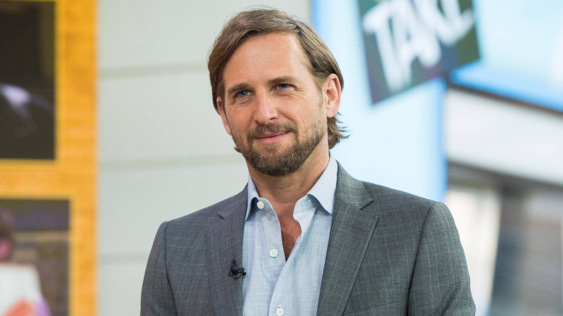 Celebrity 桌布 壁紙 — 一位留著鬍鬚、棕色頭髮的男人，穿著灰色西裝和淺藍色襯衫微笑。