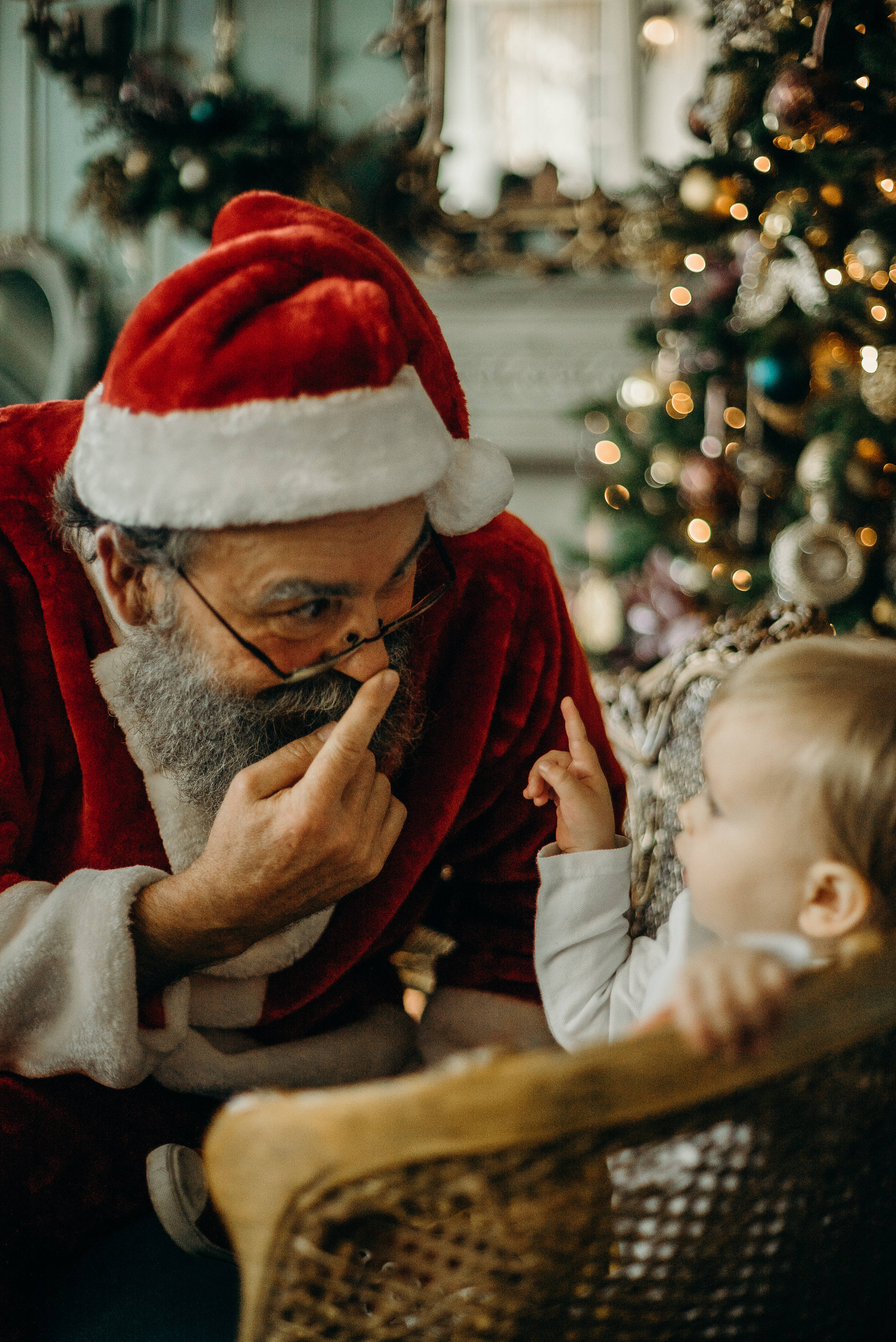 Santa Claus 桌布 壁紙 — 一位留著鬍鬚、身穿紅色西裝的聖誕老人輕輕觸摸自己的鼻子，而一個嬰兒在裝飾好的聖誕樹旁好奇地指著他。