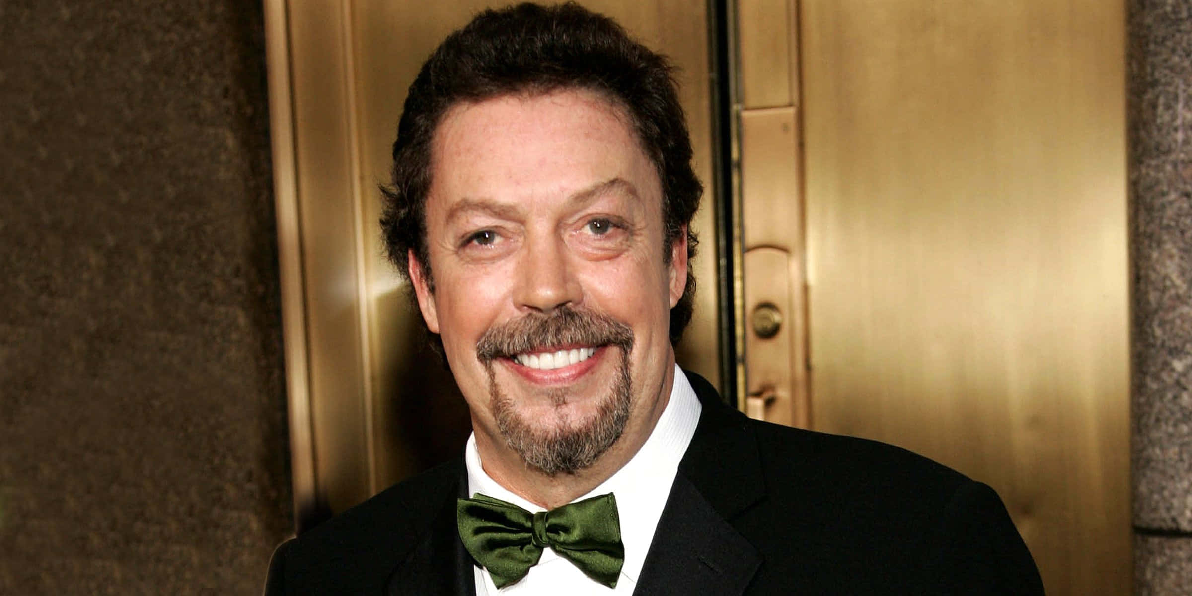 Tim Curry 桌布 壁紙 — 一位留著鬍鬚和山羊鬍的笑臉男士，身穿黑色燕尾服和綠色領結，站在金色門前。