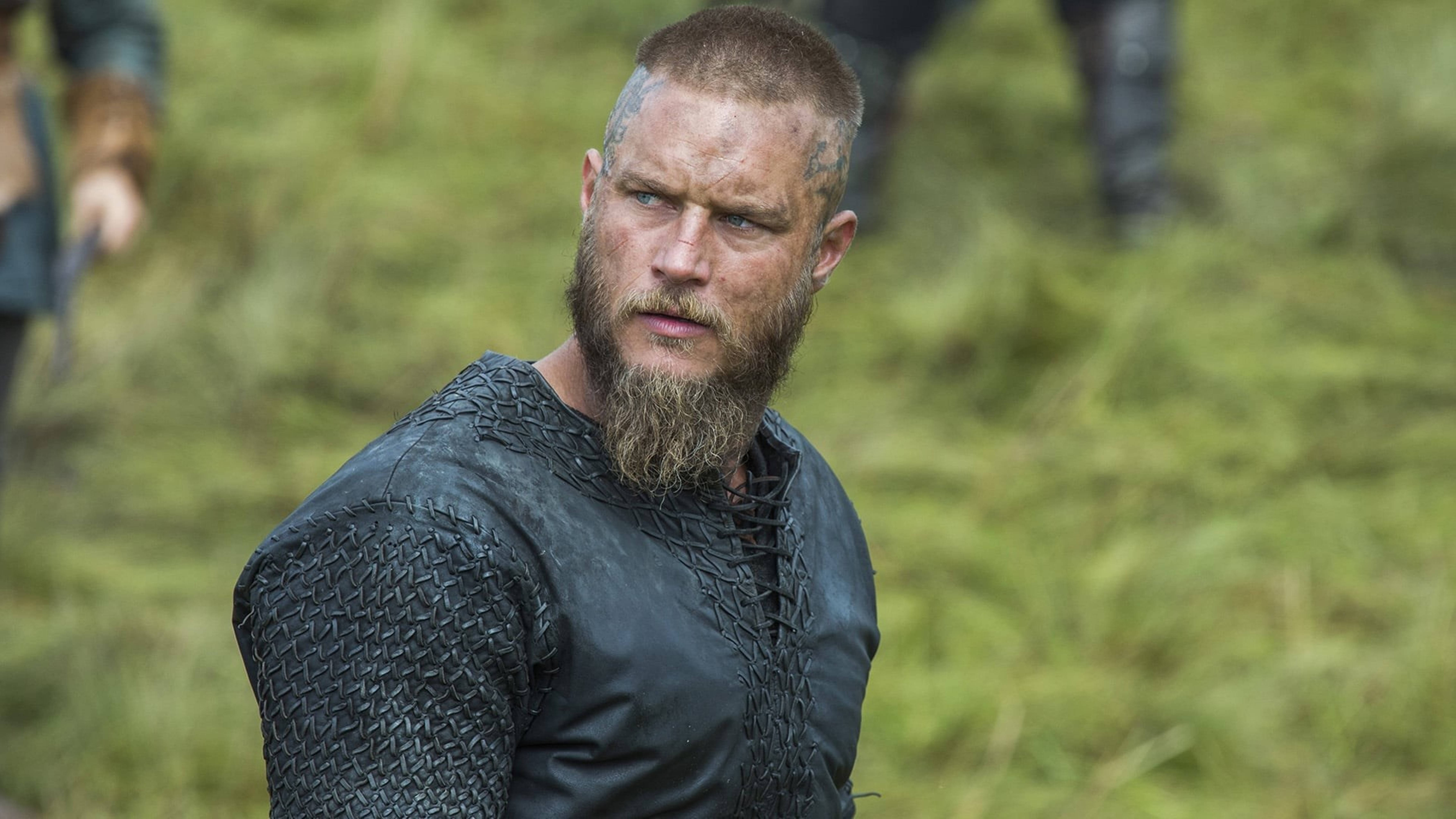 Vikings 桌布 壁紙 — 一位留著鬍鬚的維京戰士身穿鎖子甲，站在草地上，表情嚴肅。