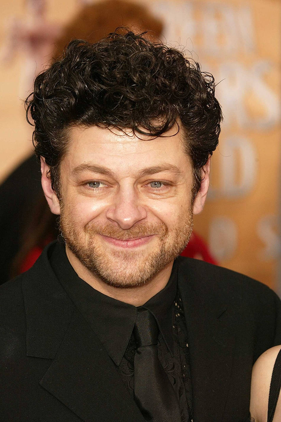 actor 桌布 壁紙 — 一位留著黑色捲髮和鬍鬚的男士在頒獎典禮上穿著黑色西裝溫暖微笑的特寫肖像。