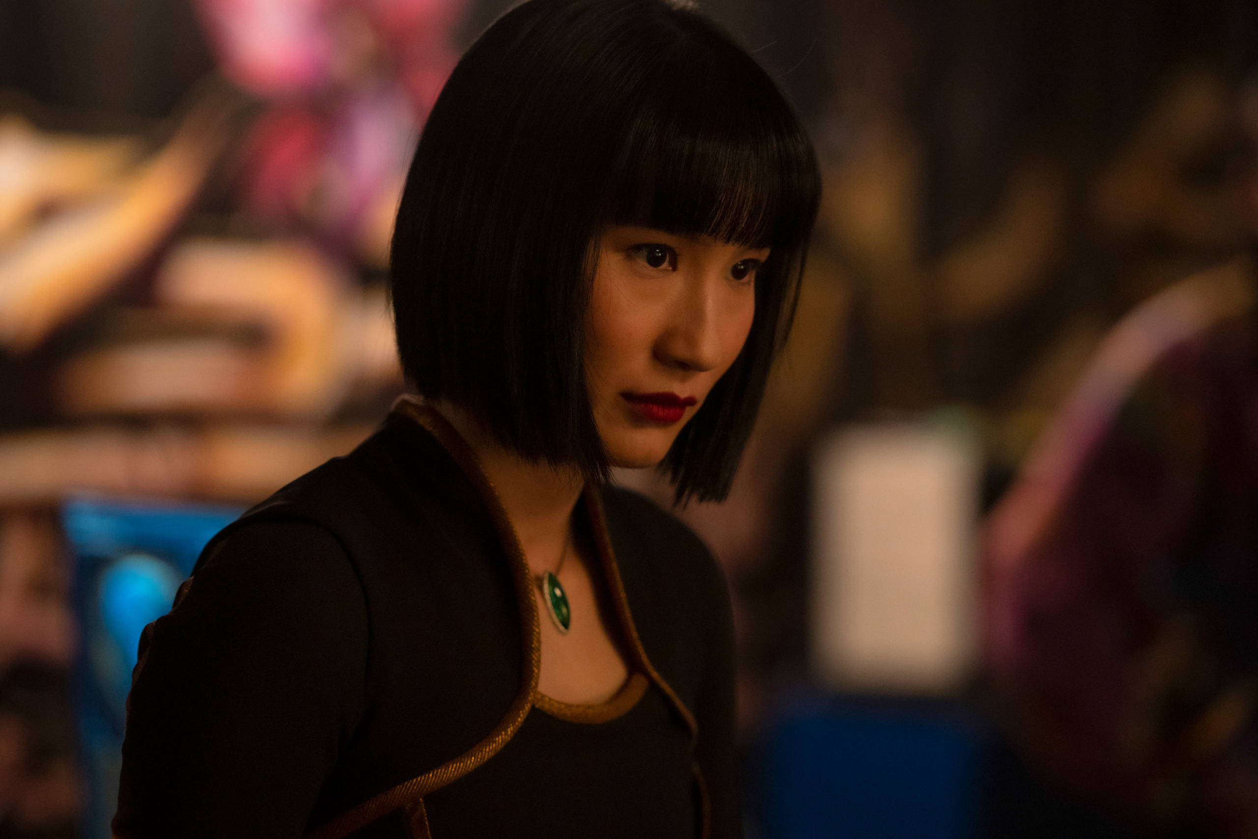 Shang 桌布 壁紙 — 一位留著黑色短鮑伯頭、紅唇的女性在昏暗、色彩繽紛的模糊背景中專注地凝視。