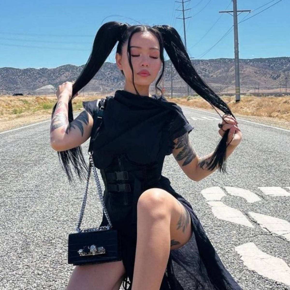 Bella Poarch 桌布 壁紙 — 一位留著黑色長辮子、有刺青的女性坐在沙漠公路上，在清澈的藍天下抓著頭髮。