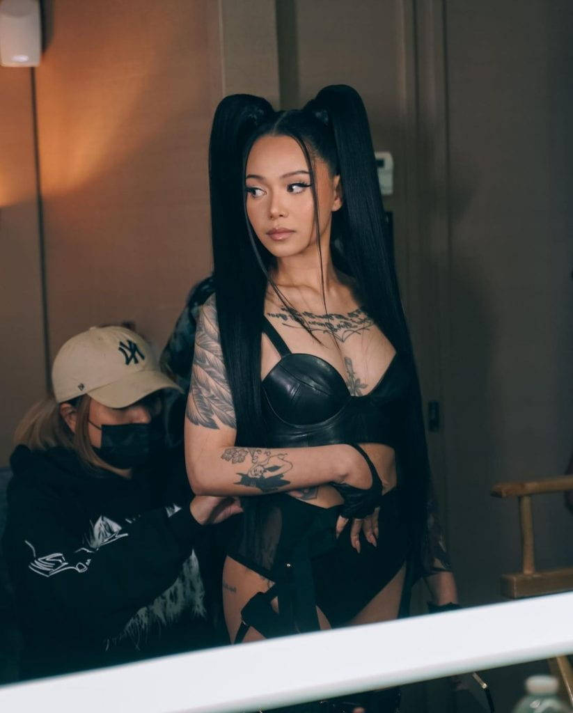 Famous 桌布 壁紙 — 一位留著黑色長辮子、有刺青的女性，穿著黑色皮革服裝，在室內與一名工作人員站在一起。