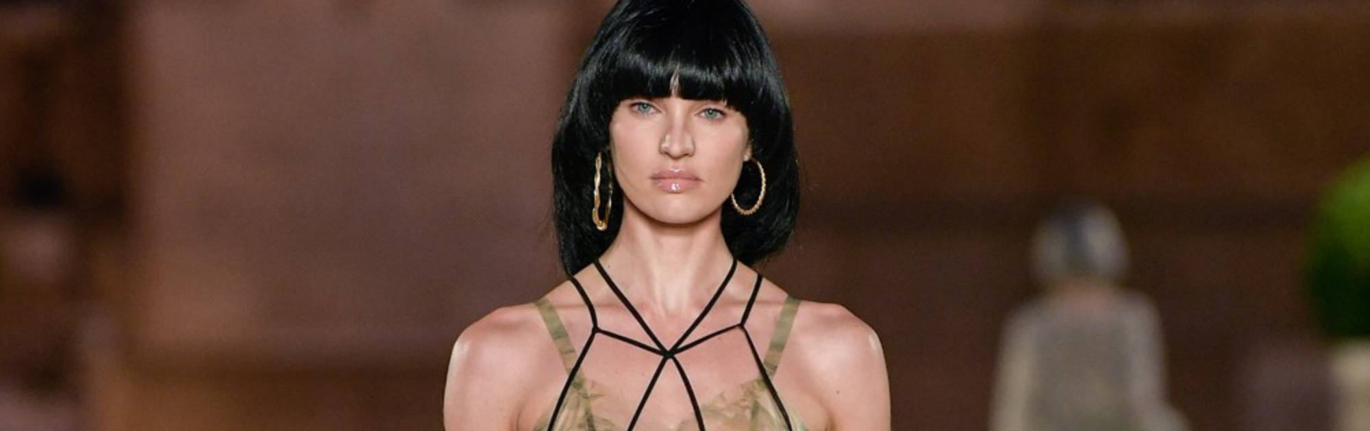 Candice 桌布 壁紙 — 一位留著黑色鮑伯頭、戴著金色圓環耳環的模特兒，自信地穿著綠色吊帶裙走在天橋上。