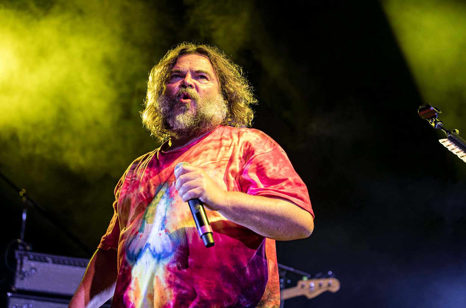 jack black 桌布 壁紙 — 一位留長鬍鬚的長髮男子在充滿活力的黃色舞台燈光下熱情地對著麥克風唱歌。