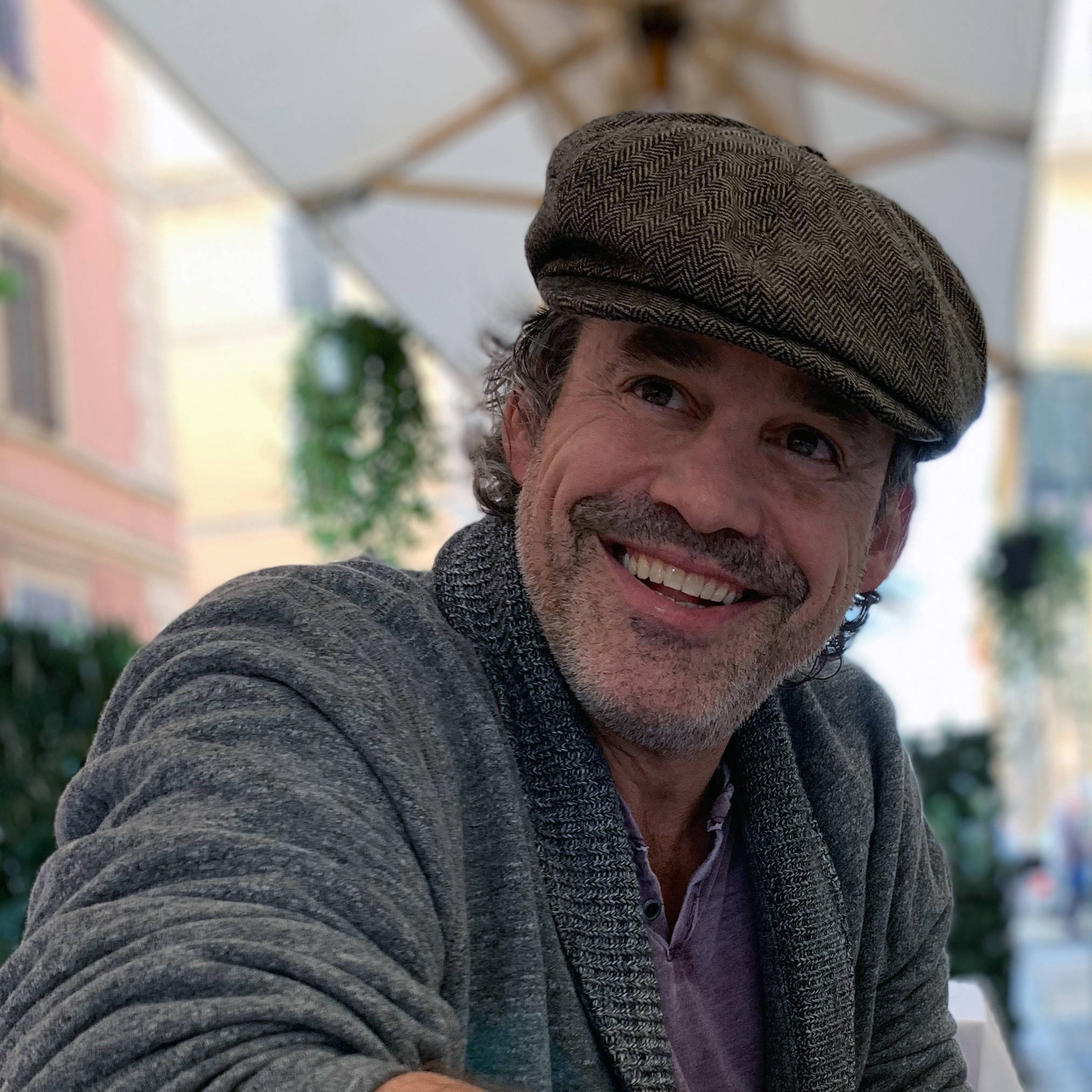 Nicholas Brendon 桌布 壁紙 — 一位留鬍子、戴人字紋帽的開朗男士，在白色庭院傘下溫暖地微笑。