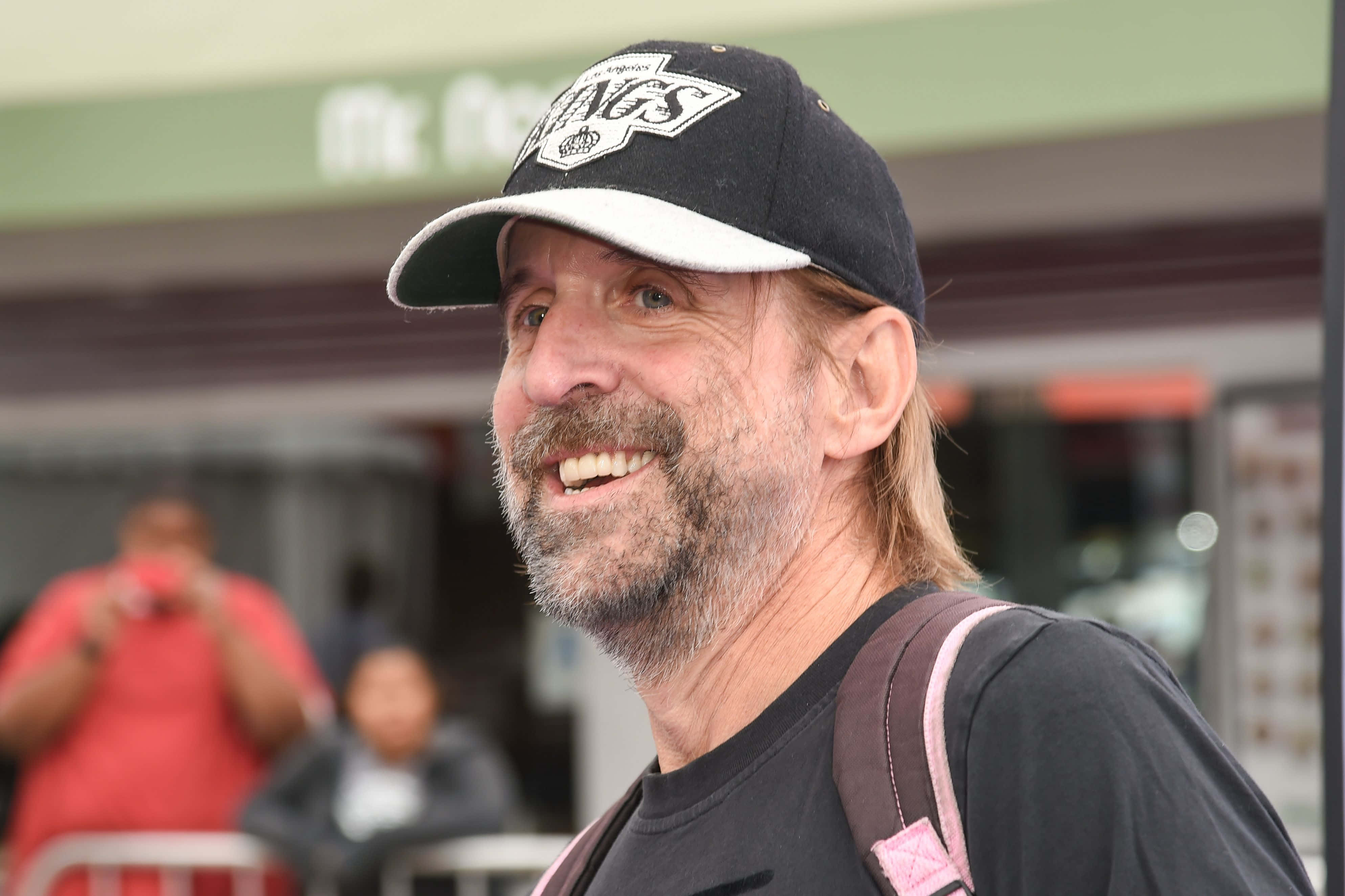 Peter Stormare 桌布 壁紙 — 一位留鬍子、戴帽子的開朗中年男子，背著背包在戶外溫暖地微笑。