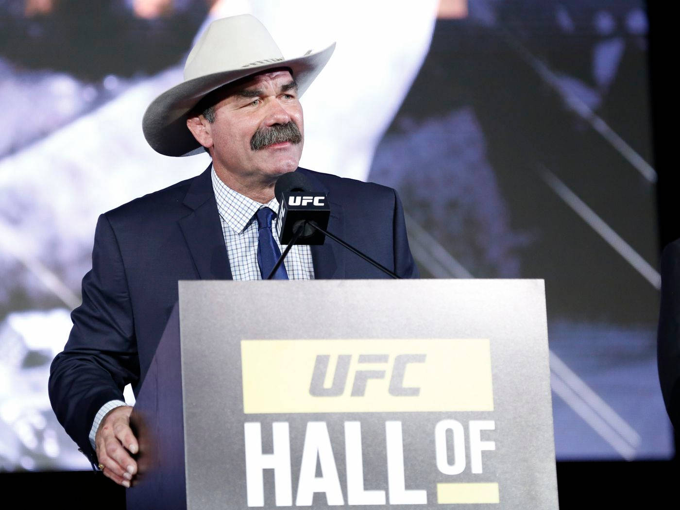 UFC 桌布 壁紙 — 一位留鬍子、戴牛仔帽的男子在明亮的舞台燈光下，於UFC名人堂的講台上發言。