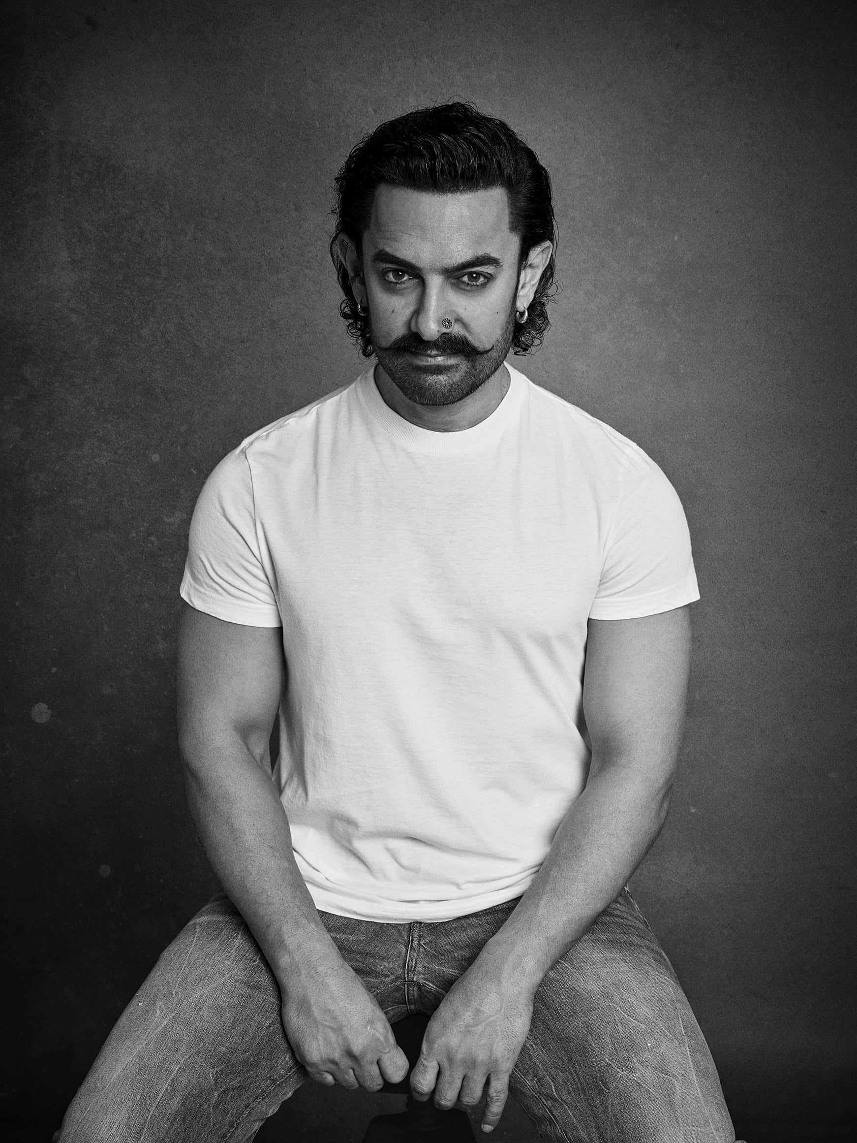 Aamir Khan 桌布 壁紙 — 一位留鬍子、戴耳環的男子自信地坐在黑白工作室肖像中，背景是帶紋理的灰色。