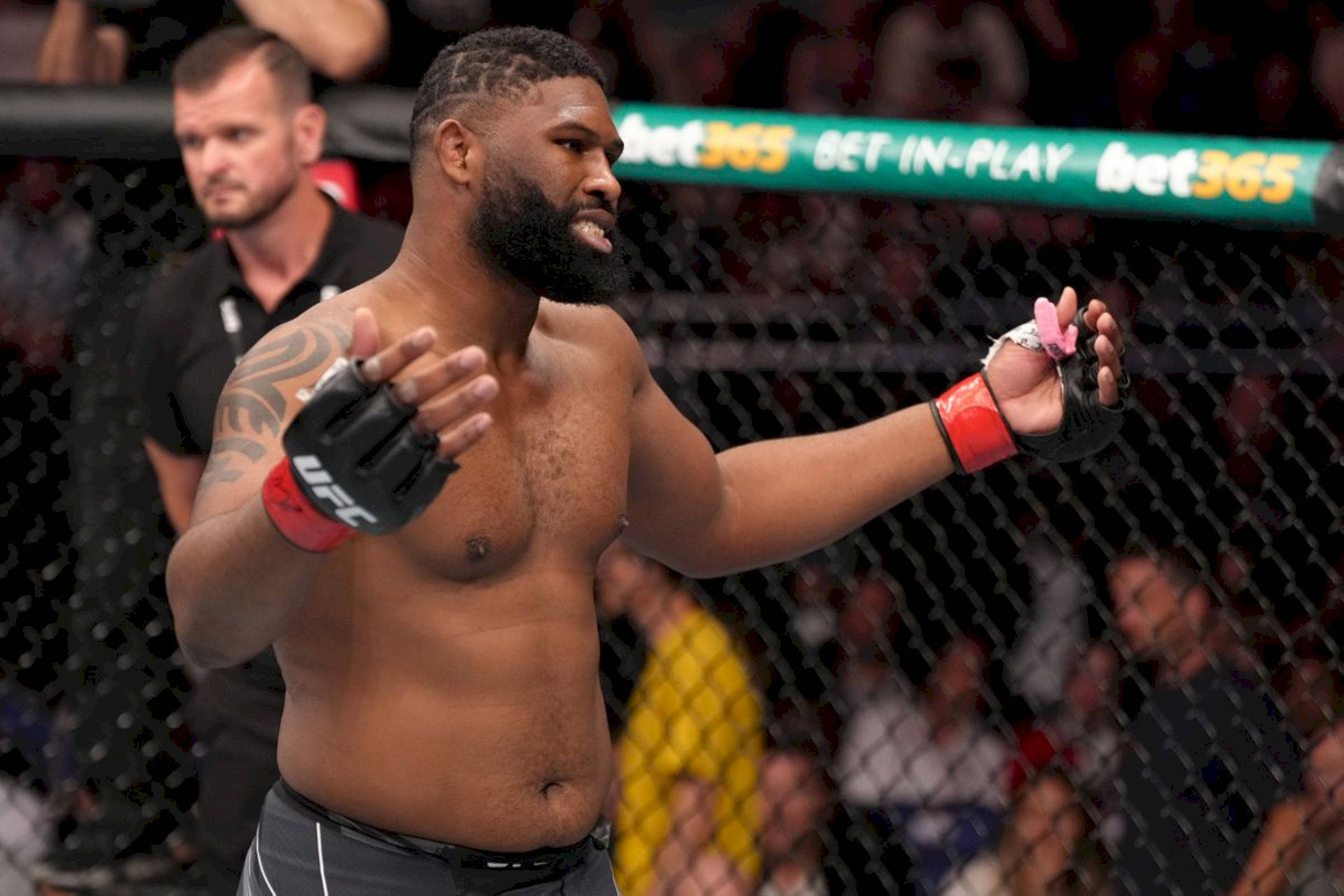 UFC 桌布 壁紙 — 一位留鬍子、手臂有刺青的肌肉發達鬥士，在UFC八角籠內以裁判和人群為背景，做出勝利的手勢。