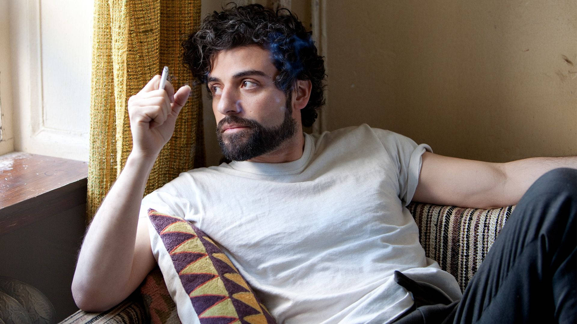 inside llewyn davis 桌布 壁紙 — 一位留鬍子、捲髮的男人，放鬆地坐在有圖案的沙發上，在黃色窗簾的窗邊拿著香煙。