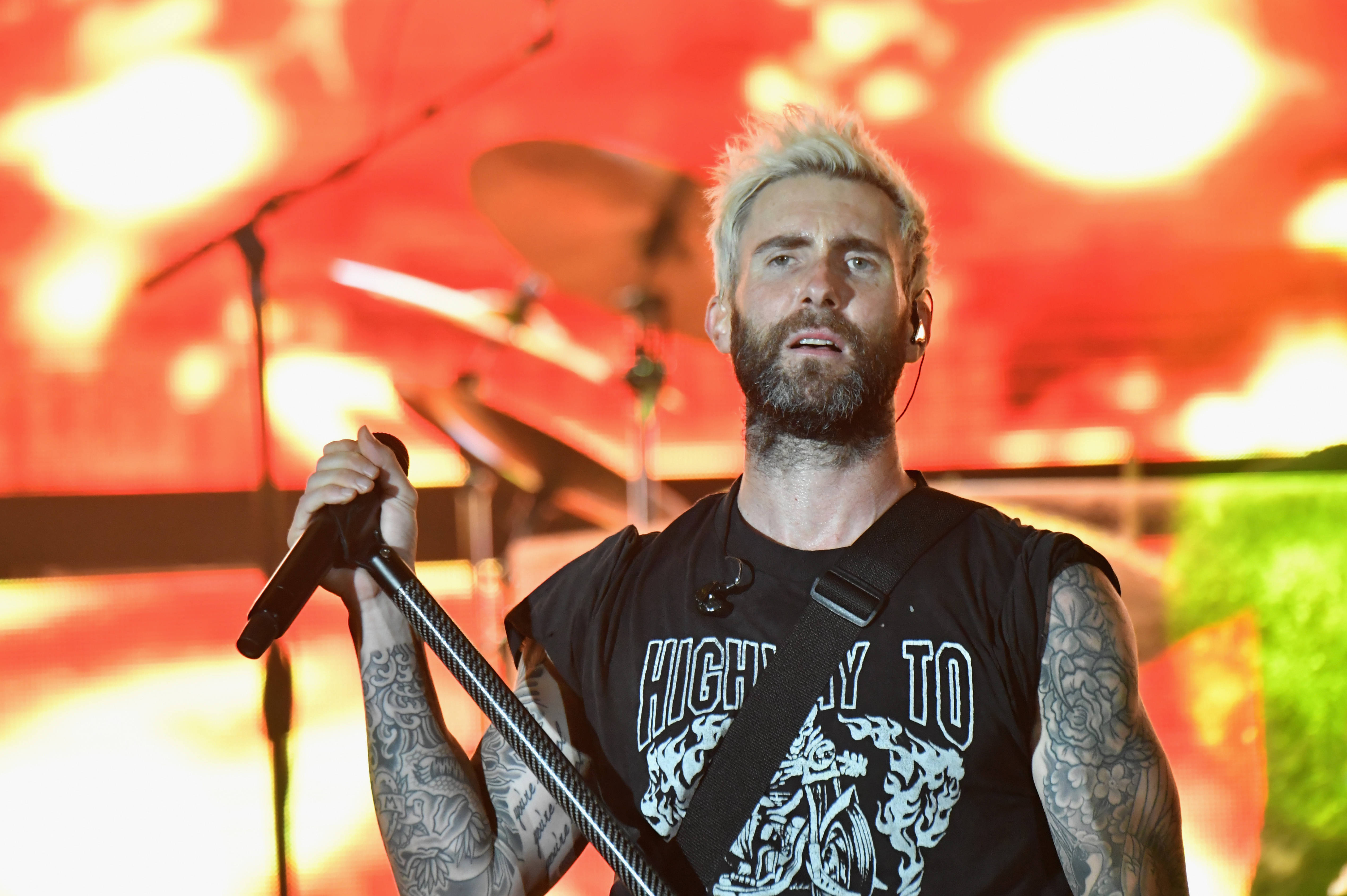 Adam Levine 桌布 壁紙 — 一位留鬍子、有刺青、漂白頭髮的歌手在舞台上表演，在火紅和橙色的燈光下拿著麥克風。