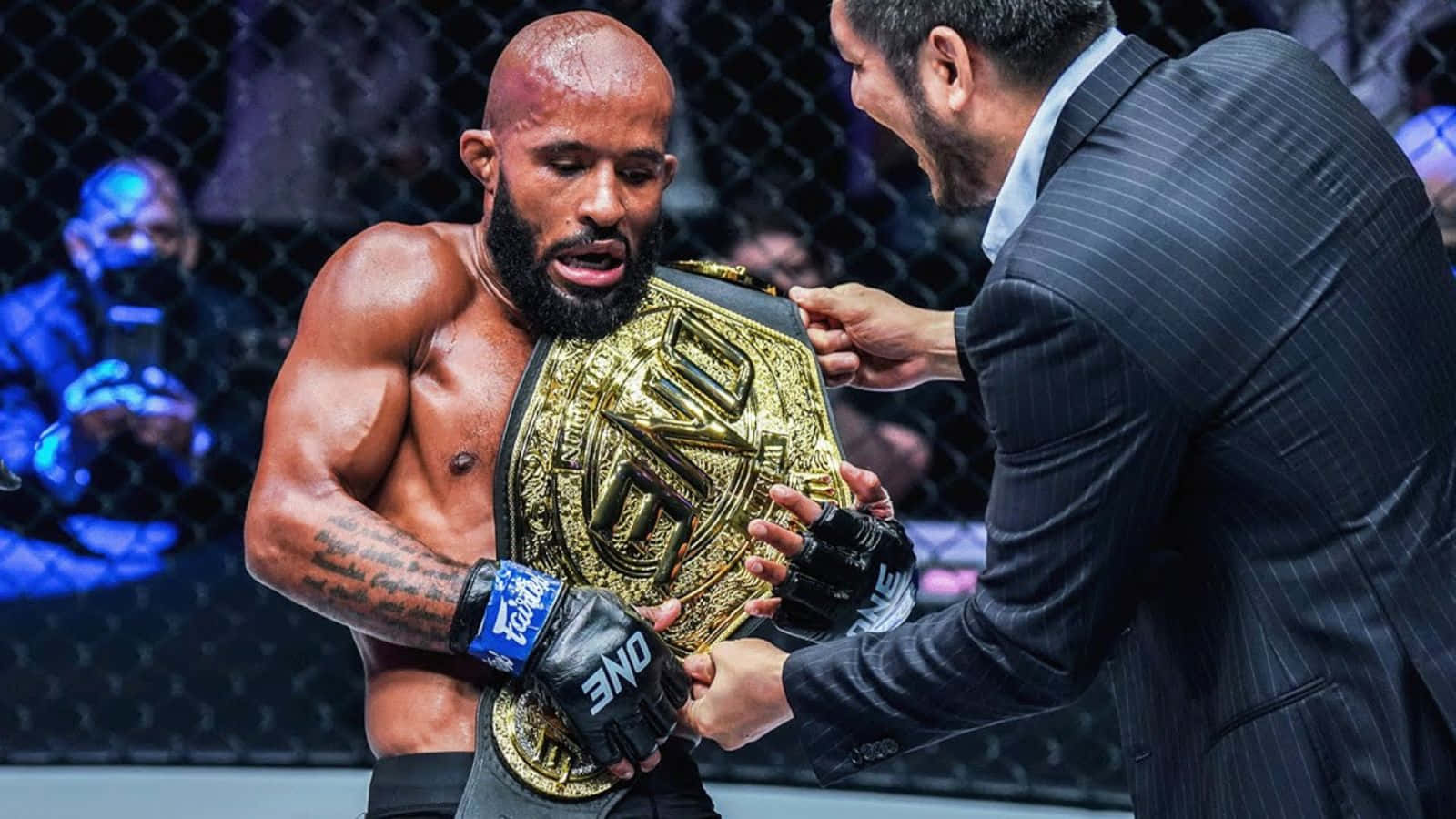 運動 桌布 壁紙 — 一位留鬍子、有刺青的勝利MMA拳手在八角籠內從一位穿西裝的男人手中接過金色冠軍腰帶。