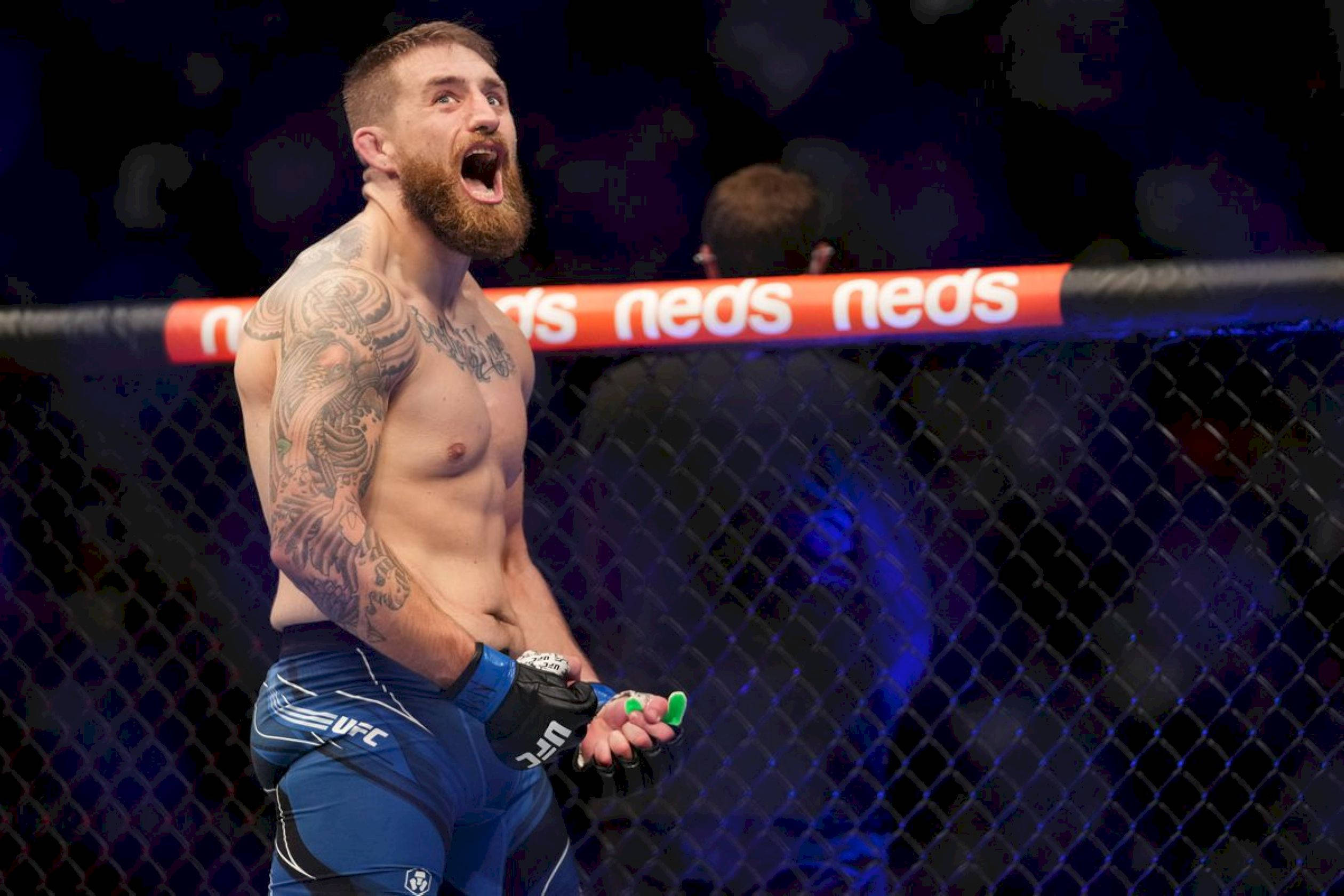 UFC 桌布 壁紙 — 一位留鬍子、有刺青的拳手在明亮的場館燈光下，於UFC八角籠內勝利地咆哮。