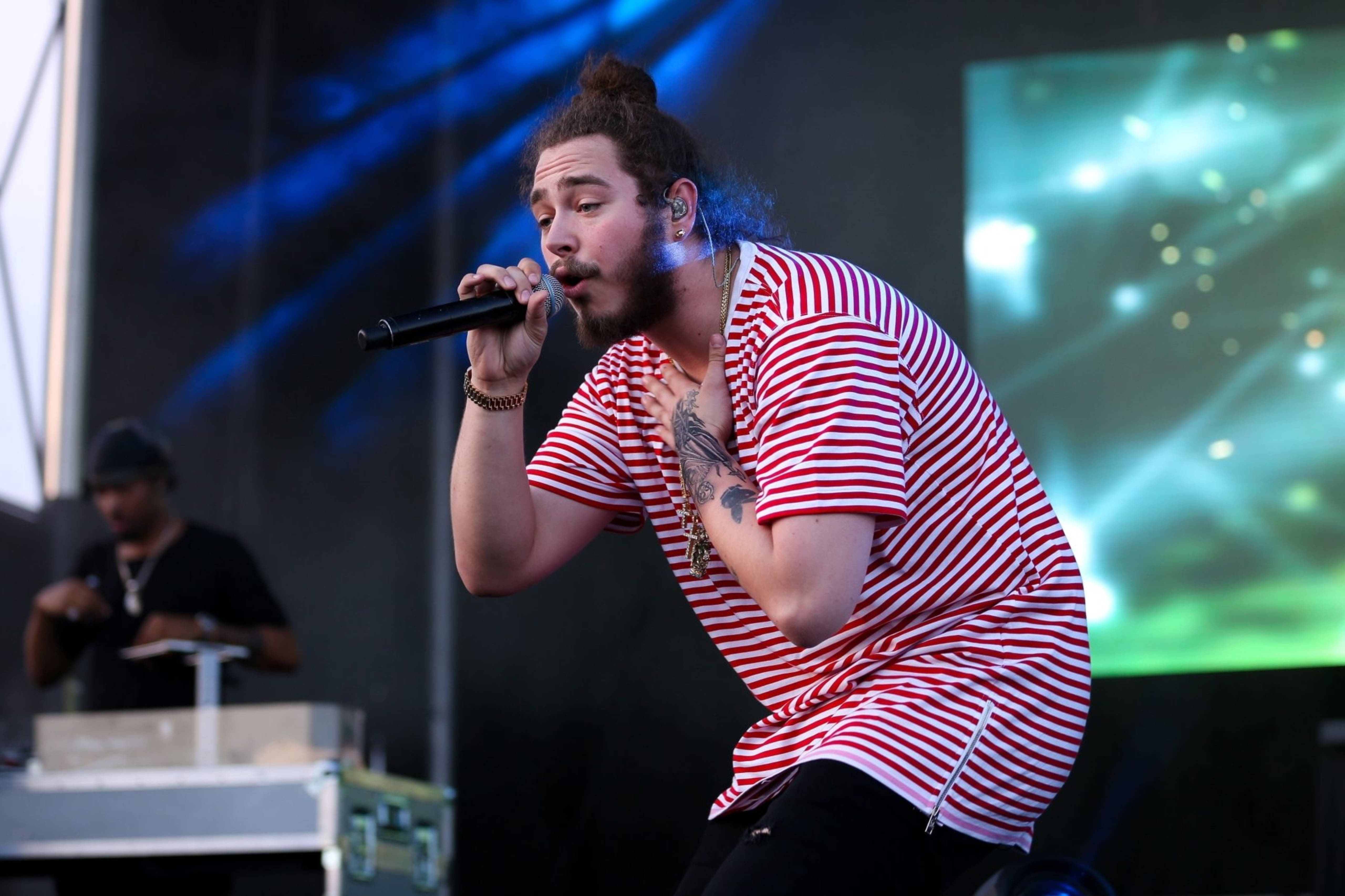Post Malone 桌布 壁紙 — 一位留鬍子、束髮髻的男子在燈光璀璨的舞台上，對著麥克風熱情獻唱，舞台燈光呈藍色和綠色。