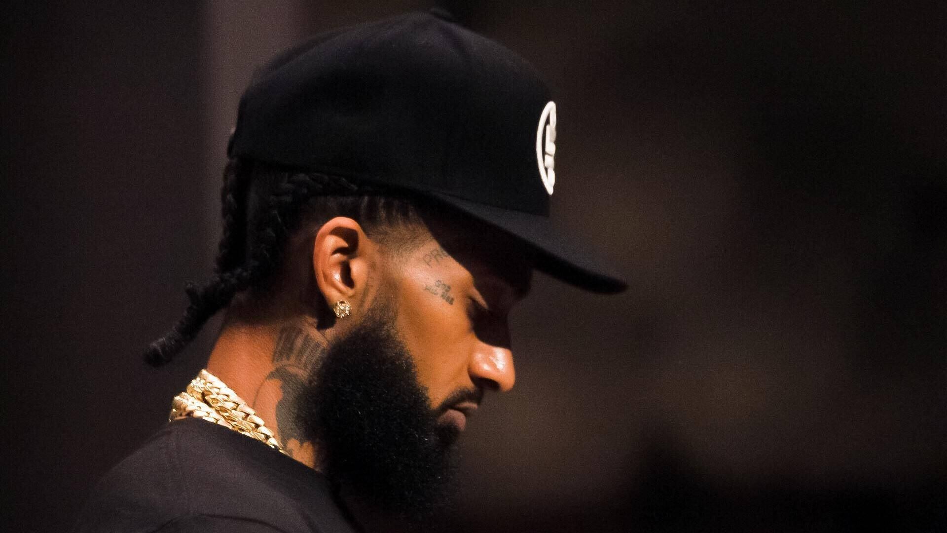 Nipsey Hussle 桌布 壁紙 — 一位留鬍子、梳辮子的男士，在昏暗的環境中戴著黑色帽子及金鏈。