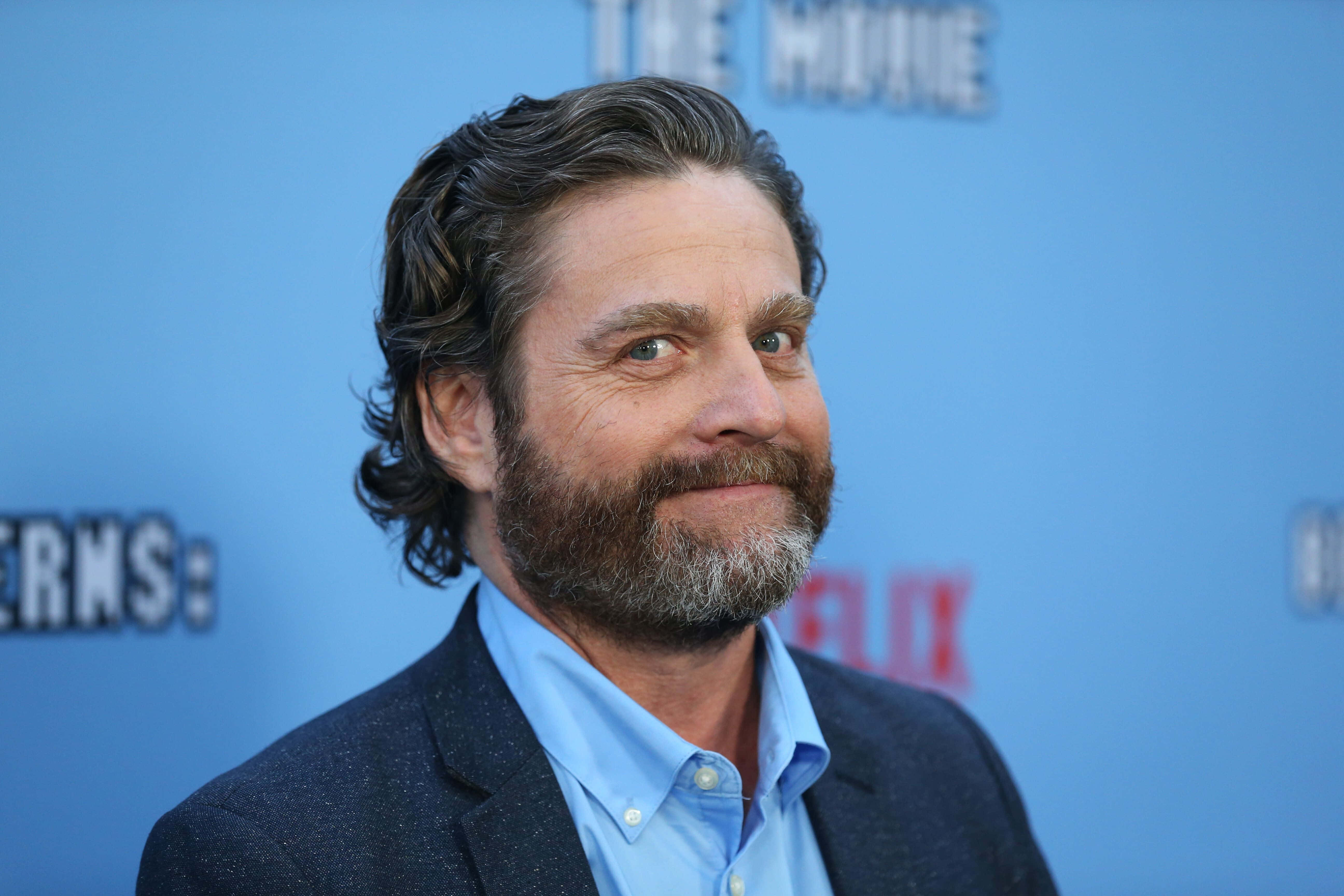 Zach Galifianakis 桌布 壁紙 — 一位留鬍子、波浪髮型的男士在藍色宣傳背景前溫暖微笑，背景上有文字。
