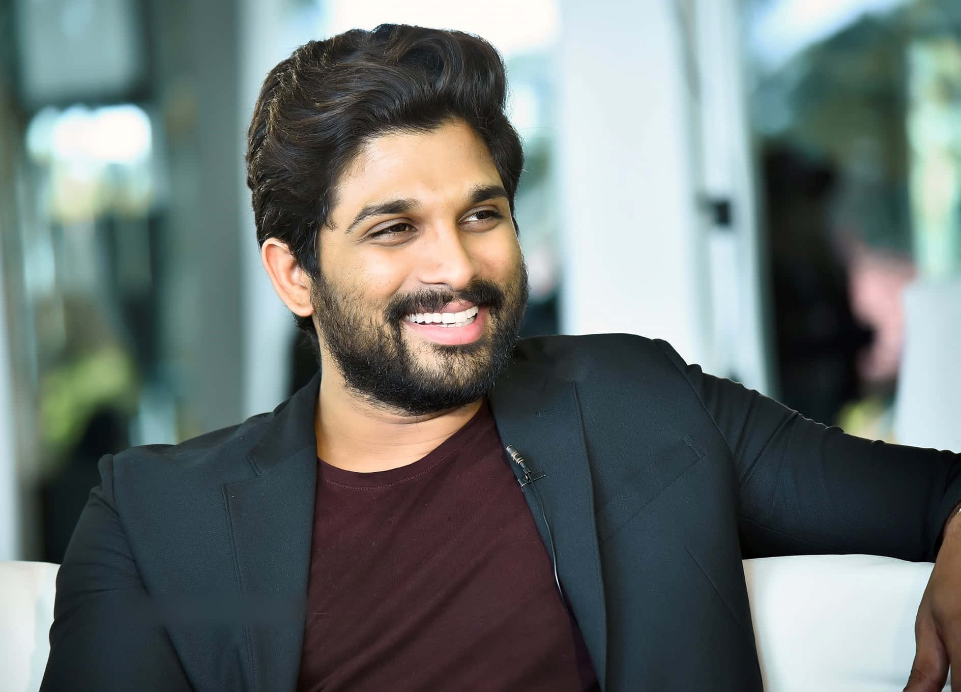 Allu Arjun 桌布 壁紙 — 一位留鬍子、深色頭髮的男士微笑著，在現代室內環境中放鬆地坐著，身穿黑色西裝外套內搭栗色襯衫。