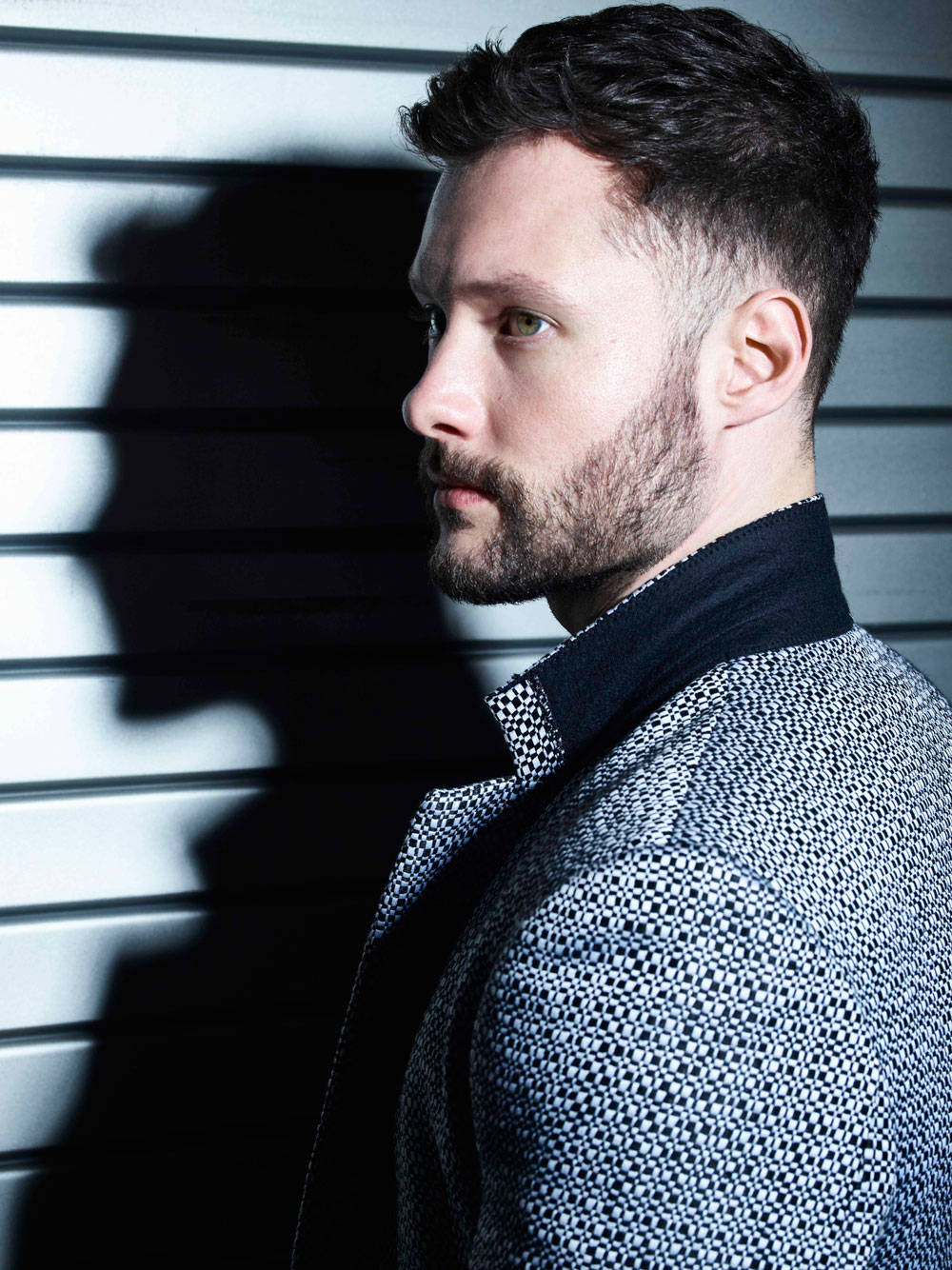 Calum Scott 桌布 壁紙 — 一位留鬍子、深色頭髮的男子穿著圖案西裝外套，側身站在條紋牆前，投射出戲劇性的陰影。
