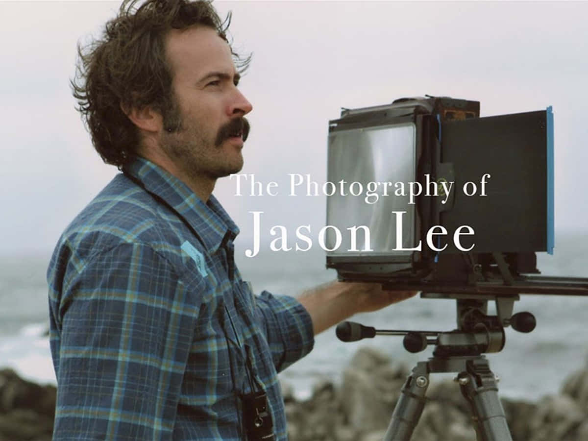Jason Lee 桌布 壁紙 — 一位留鬍子、穿著格紋襯衫的男人，在海岸背景下操作三腳架上的大畫幅相機。
