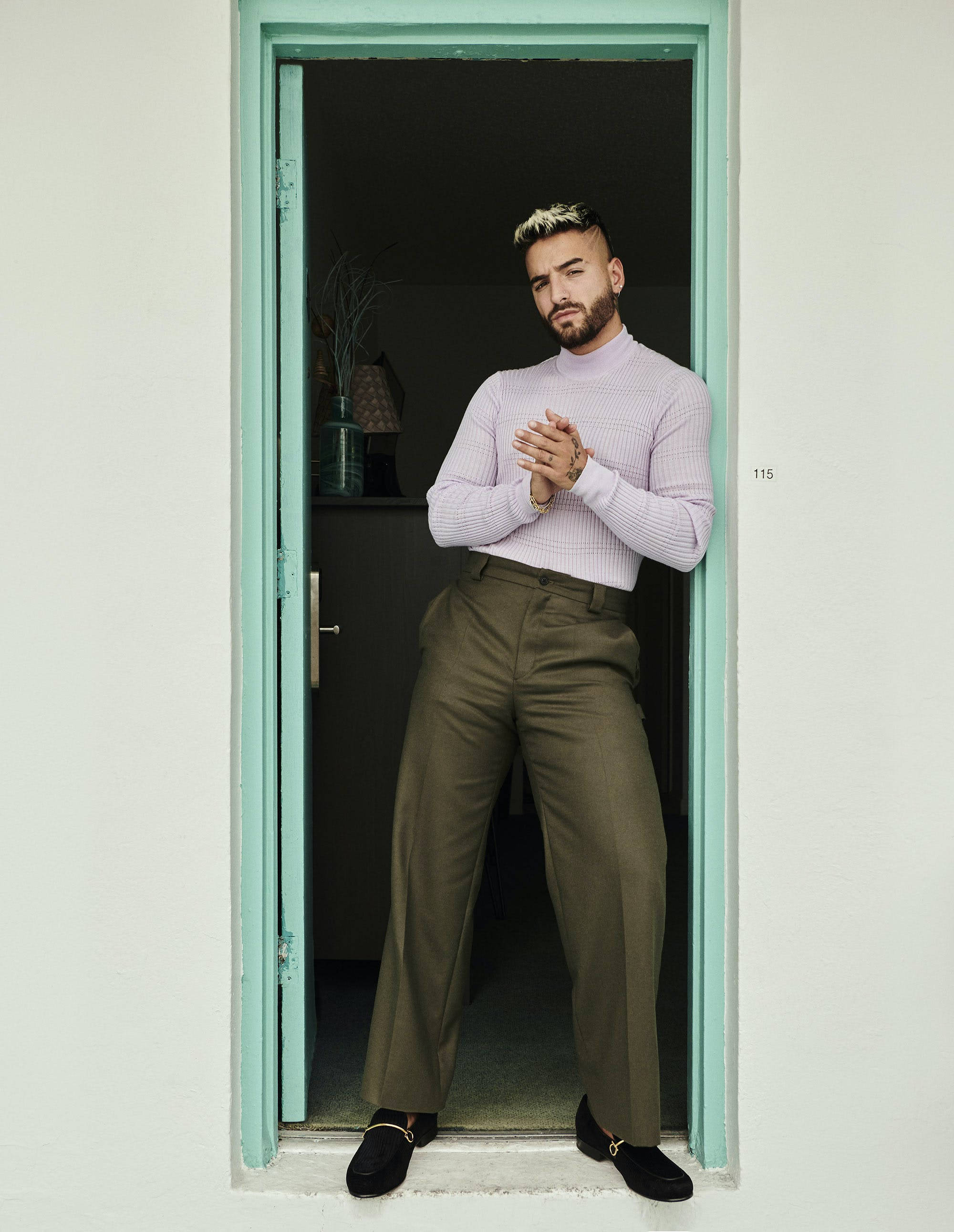 Maluma 桌布 壁紙 — 一位留鬍子、金髮的時尚男士站在薄荷綠色的門口，穿著薰衣草色高領毛衣和橄欖色褲子。