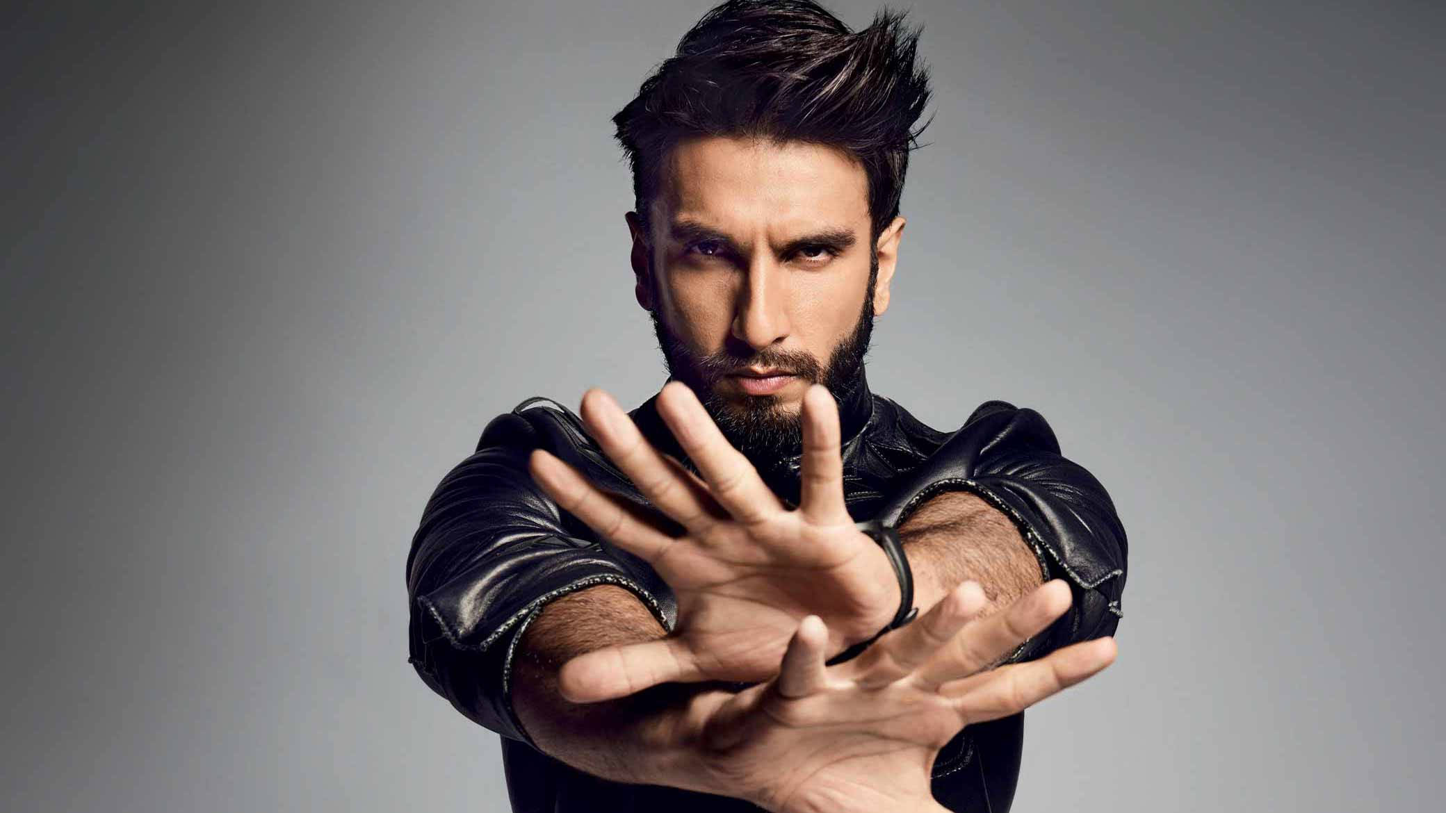 Ranveer Singh 桌布 壁紙 — 一位留鬍子、頭髮豎起的男士穿著黑色皮夾克，在灰色攝影棚背景前雙手向前擺姿勢。