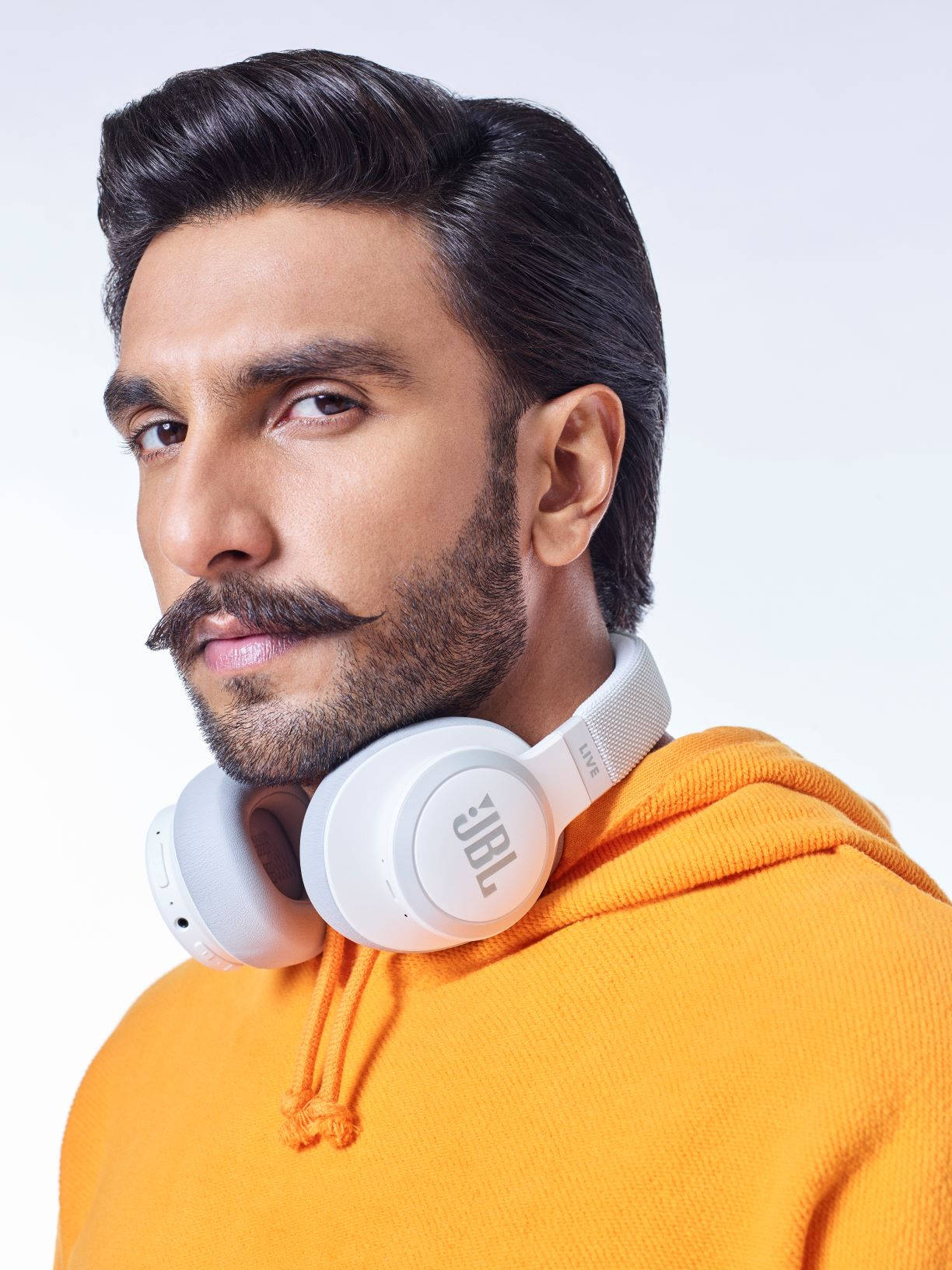 Ranveer Singh 桌布 壁紙 — 一位留鬍子、頭髮造型時尚的男士，脖子上掛著白色JBL耳機，身穿鮮橙色連帽上衣，背景是純白色。