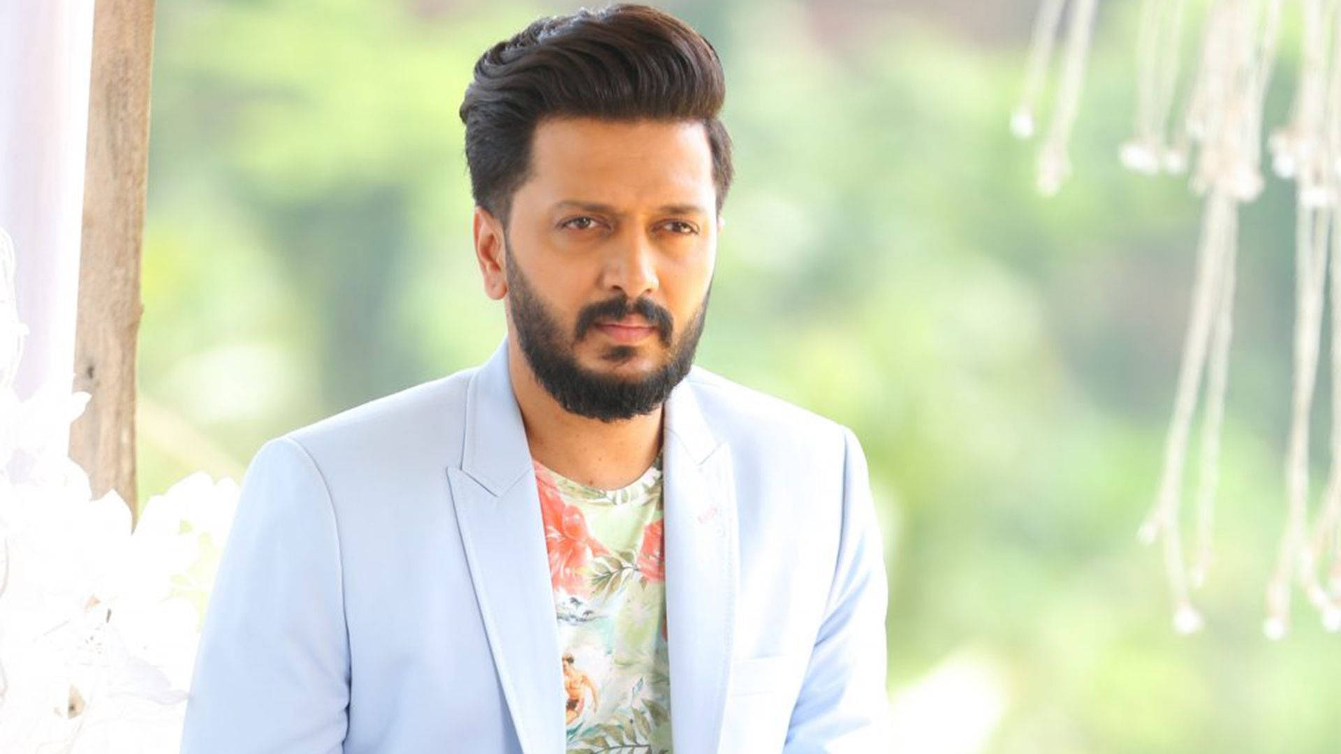riteish deshmukh 桌布 壁紙 — 一位留鬍子、頭髮造型時尚的男子，在柔和的綠色模糊背景前，穿著淺藍色西裝外套，內搭花卉圖案襯衫。