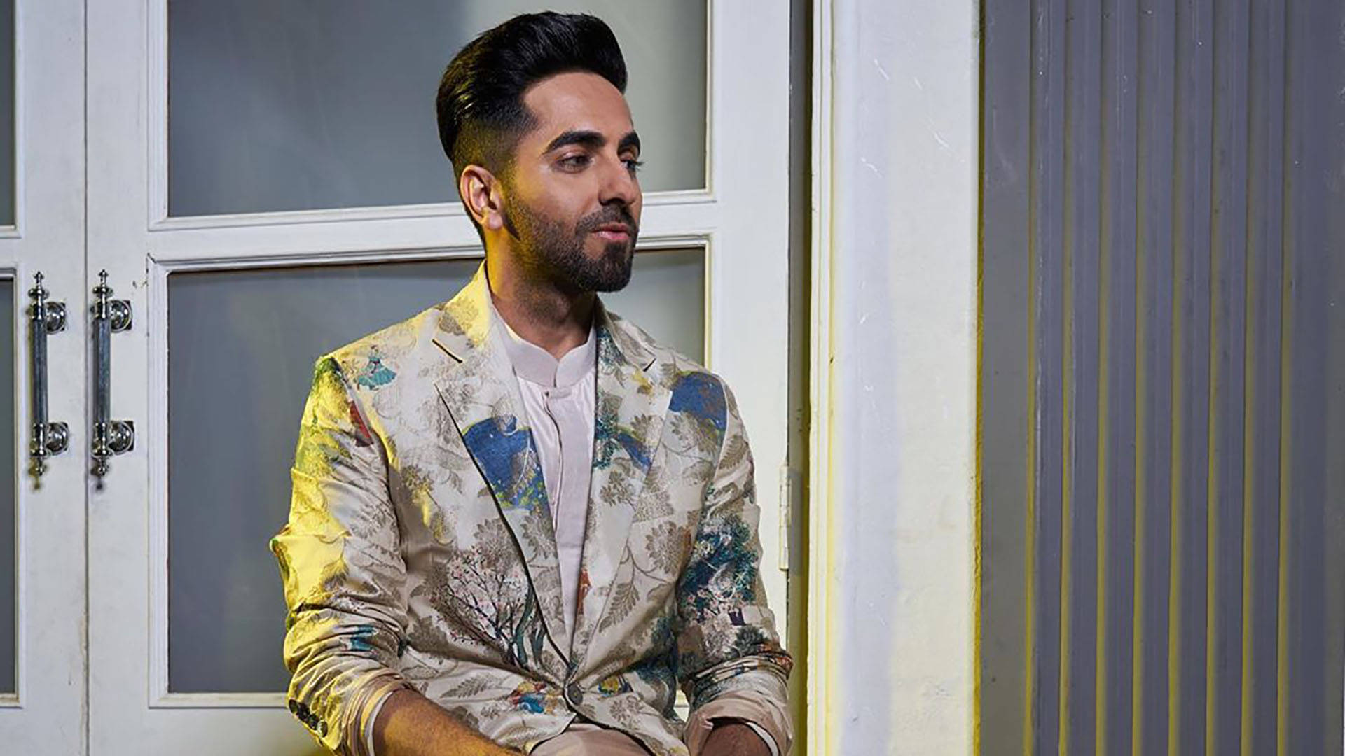 ayushmann khurrana 桌布 壁紙 — 一位留鬍子、頭髮造型時尚的男子穿著花卉圖案西裝外套，靠在一扇帶玻璃窗的白色門上。