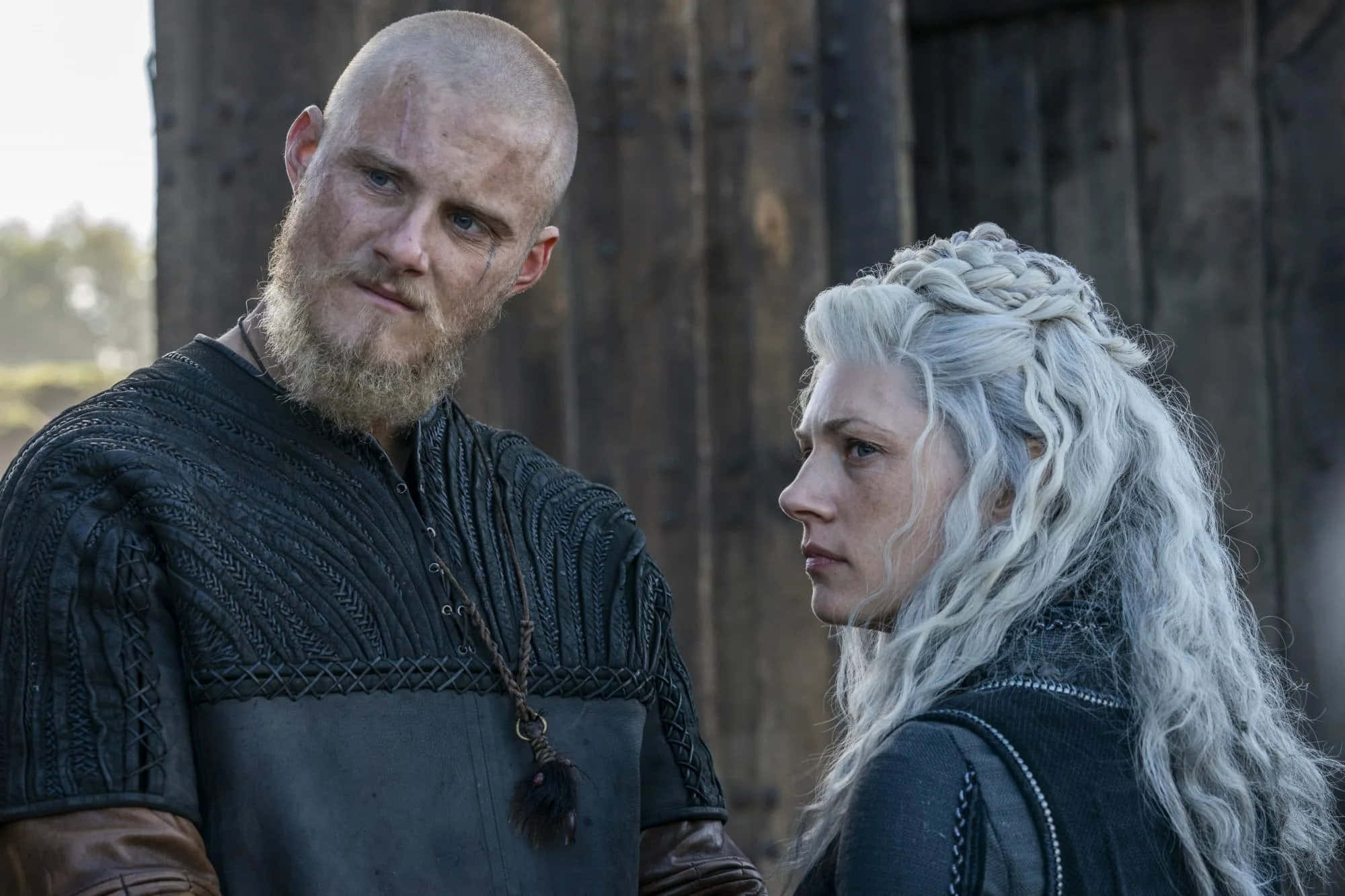 Vikings 桌布 壁紙 — 一位留鬍子光頭的維京男人站在一位辮子白髮的女人旁邊，兩人都穿著深色中世紀服裝，背景是木質的。