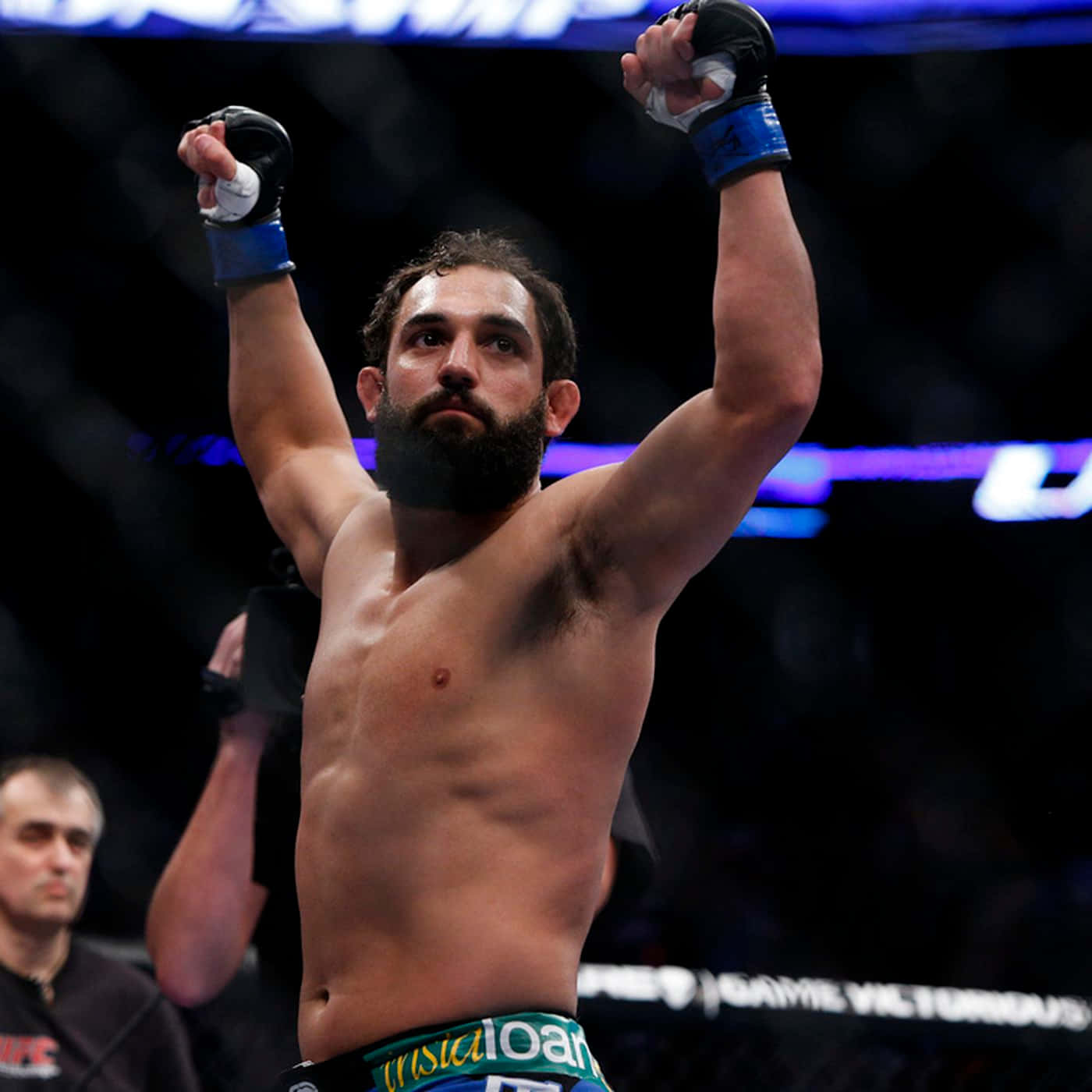 UFC 桌布 壁紙 — 一位留鬍子的MMA拳手在昏暗的八角籠中，在發光的藍光下勝利地舉起拳頭。