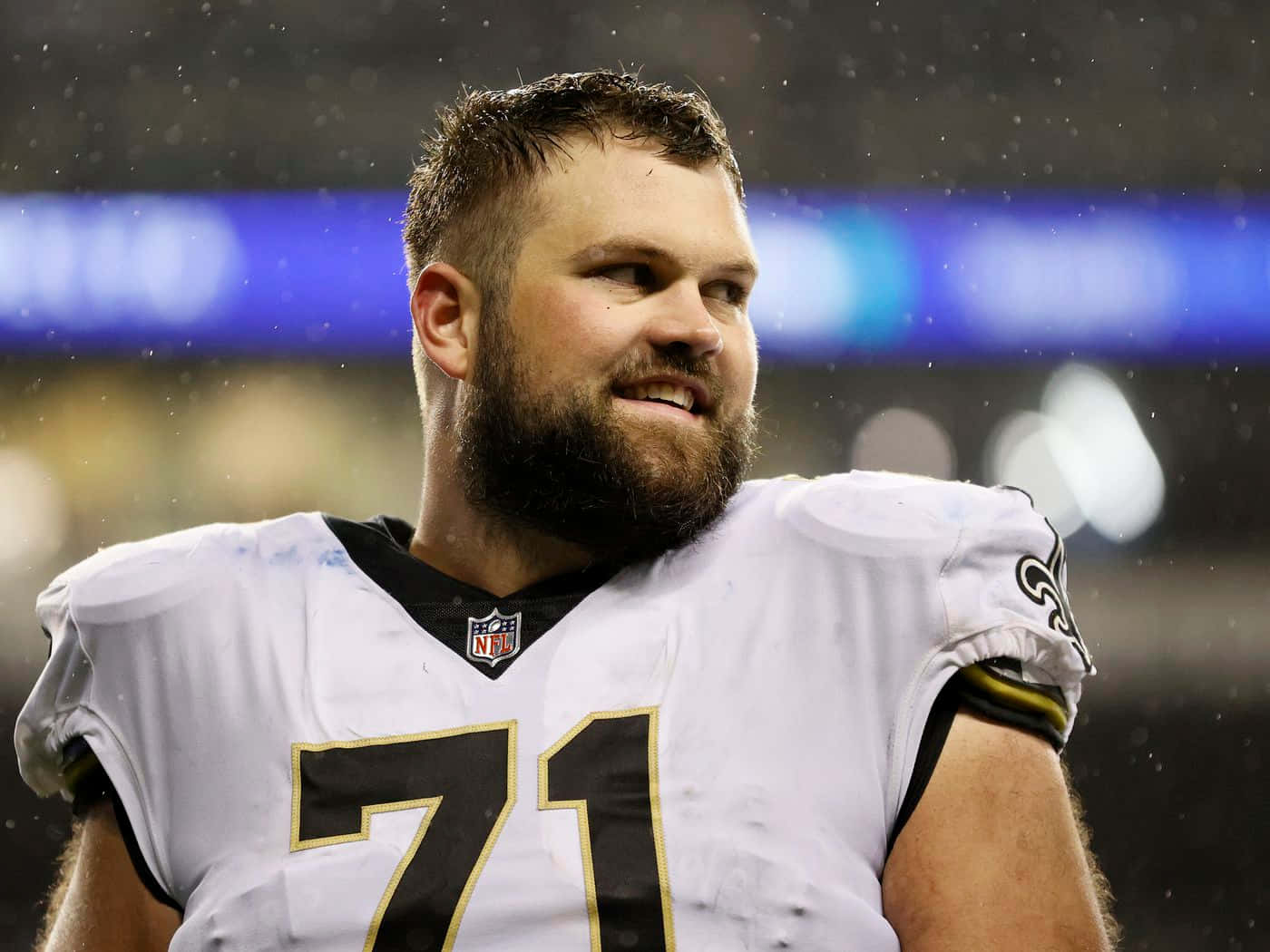 Athlete 桌布 壁紙 — 一位留鬍子的NFL球員，身穿白色球衣，號碼71，在雨中的比賽中，在體育場燈光下微笑。