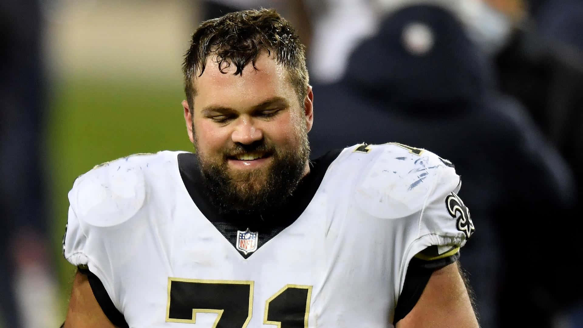 Athlete 桌布 壁紙 — 一位留鬍子的NFL球員身穿白色球衣，號碼71，在夜晚的足球場上低頭微笑。