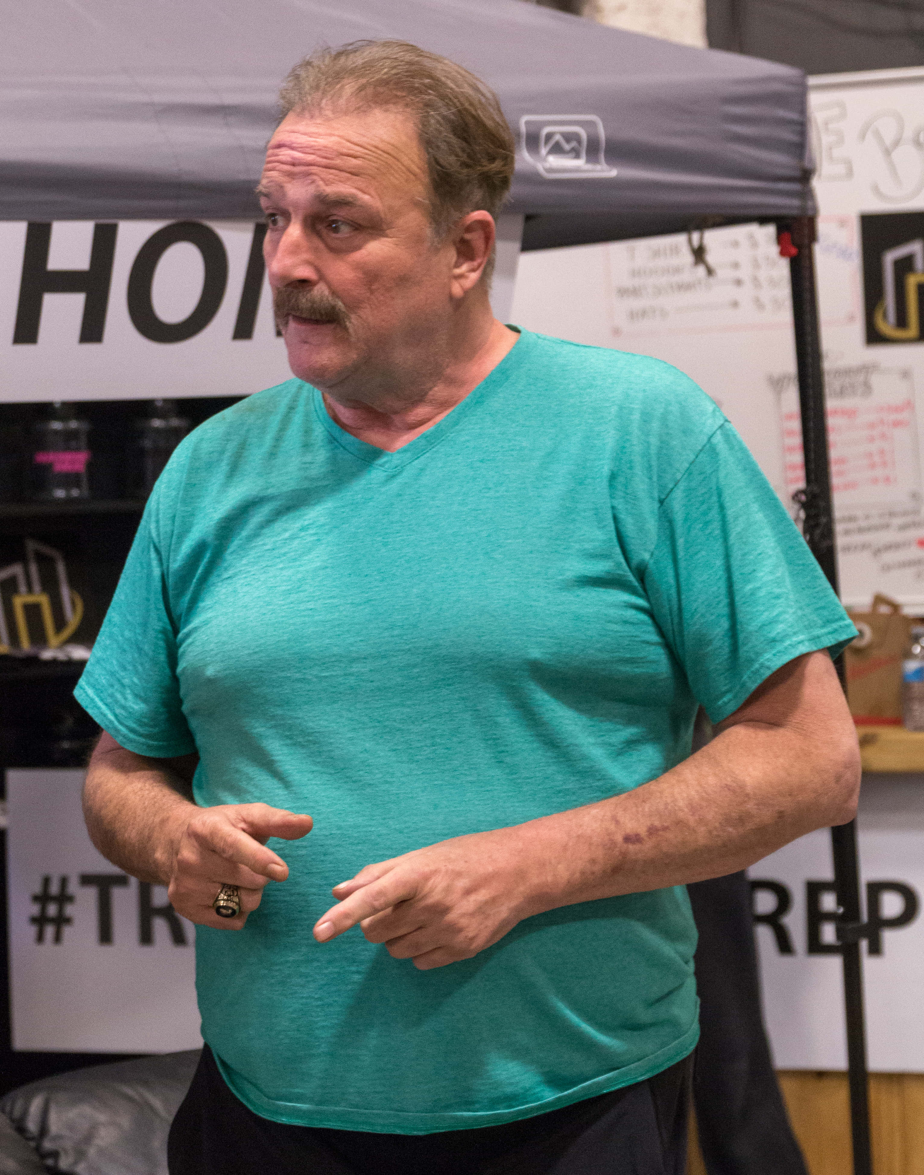 摔角 桌布 壁紙 — 一位留鬍子的中年男子穿著青綠色T恤，站在有標牌和遮陽篷的展會攤位前做手勢。