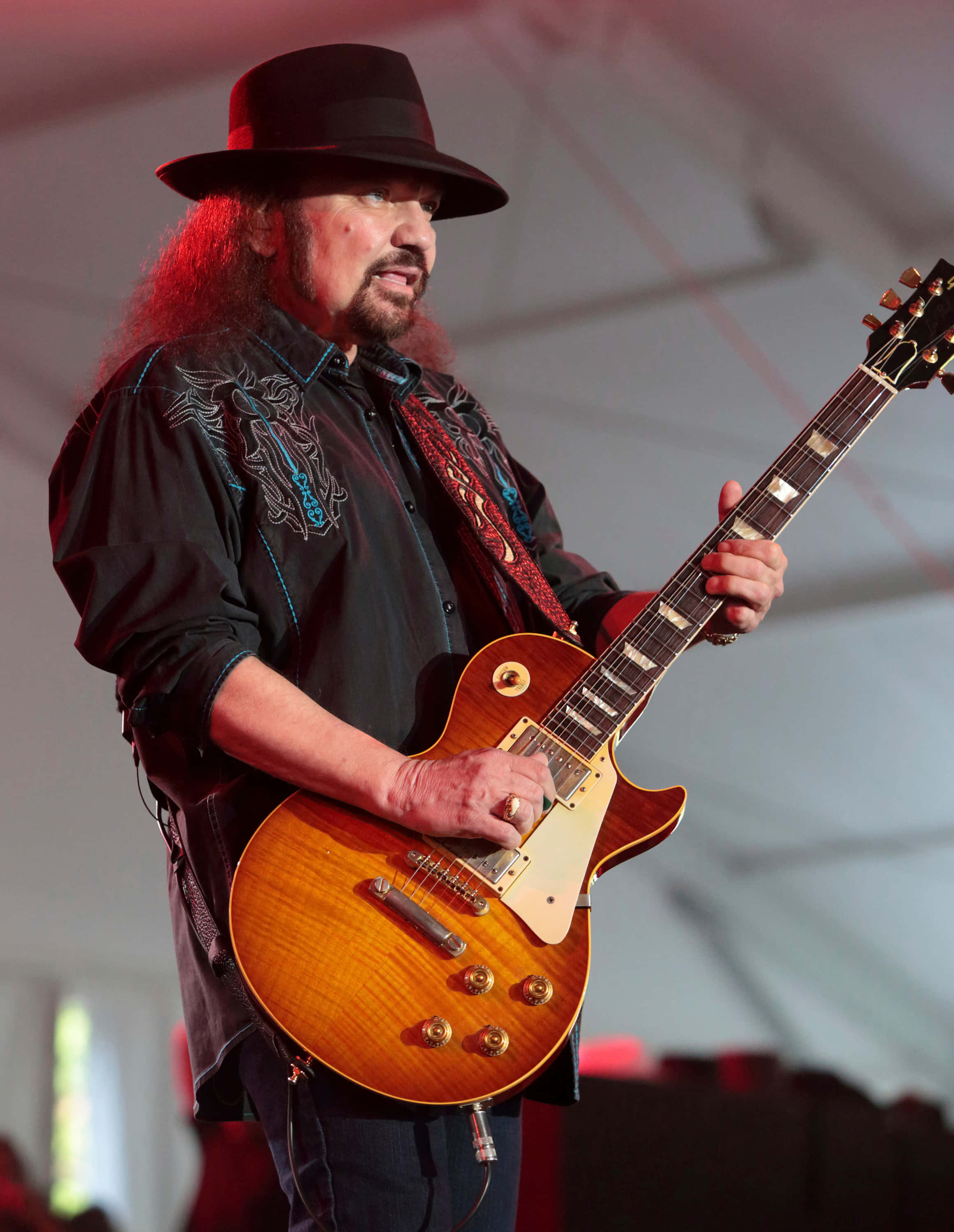 Gary Rossington 桌布 壁紙 — 一位留鬍子的吉他手，戴著黑帽和刺繡襯衫，在舞台燈光下熱情地彈奏一把旭日紋電吉他。