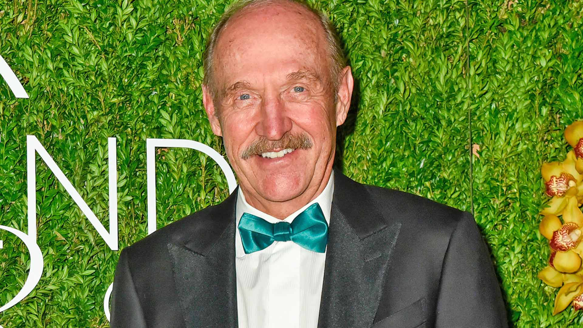 運動 桌布 壁紙 — 一位留鬍子的微笑年長男士，穿著黑色燕尾服和青綠色領結，背景是茂密的綠色樹籬。