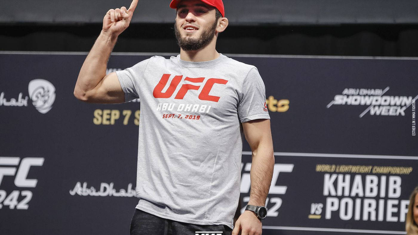 UFC 桌布 壁紙 — 一位留鬍子的戰士，戴著紅色帽子，穿著灰色UFC T恤，在稱重活動中自信地微笑並向上指。