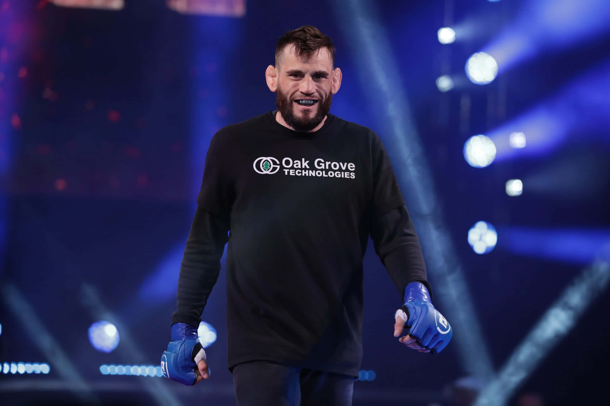 UFC 桌布 壁紙 — 一位留鬍子的戰士，戴著藍色手套，穿著黑色「Oak Grove Technologies」襯衫，在明亮的舞台燈光下自信地微笑。