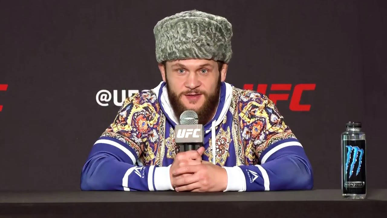 UFC 桌布 壁紙 — 一位留鬍子的戰士穿著圖案外套和毛帽，在記者會上對著UFC麥克風說話。