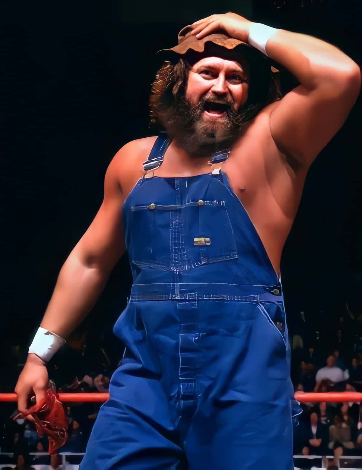 WWE 桌布 壁紙 — 一位留鬍子的摔角手，身穿藍色工裝褲和帽子，在擂台上勝利慶祝，身後是歡呼的人群。