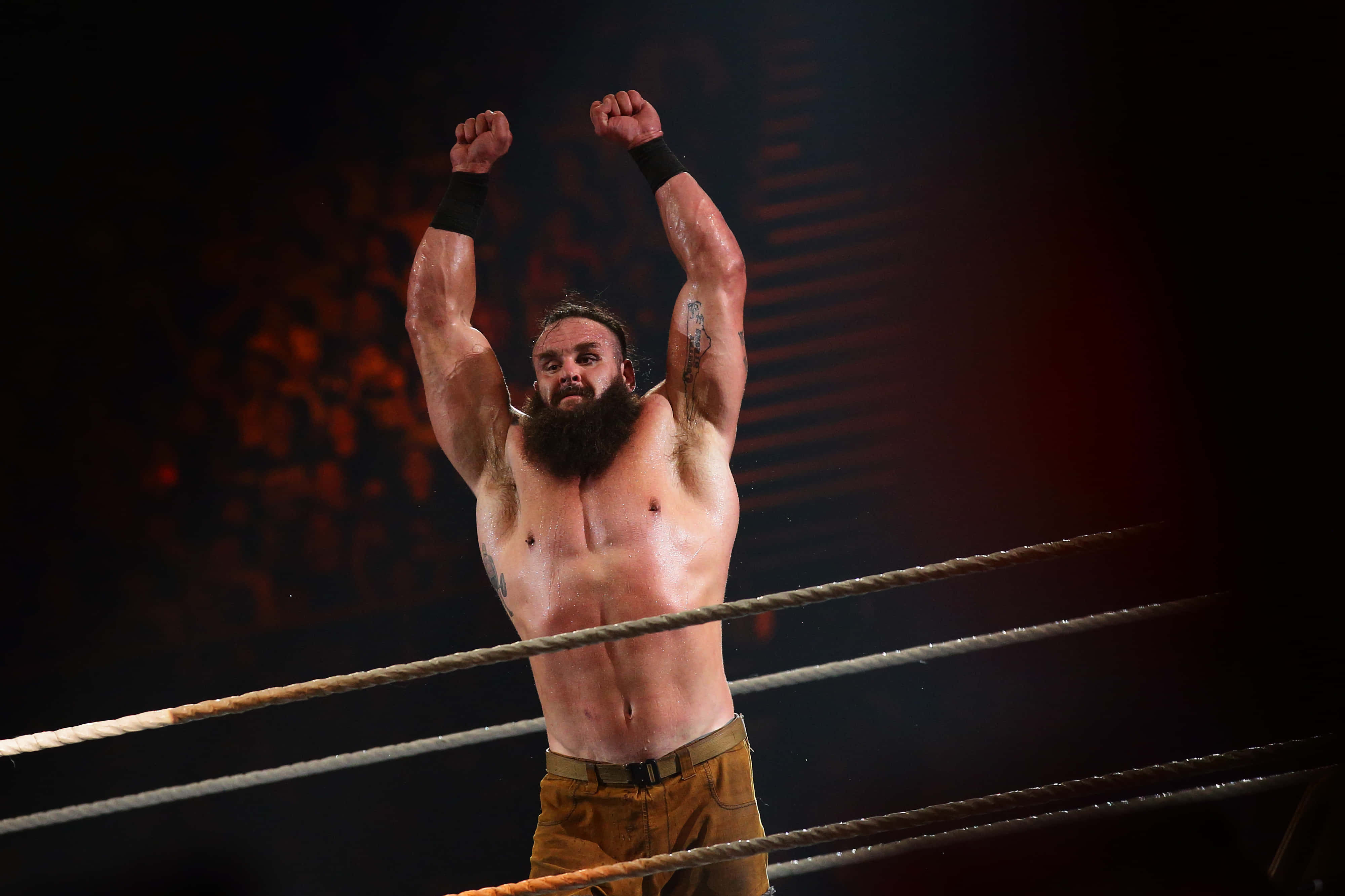 WWE 桌布 壁紙 — 一位留鬍子的摔角手在戲劇性的紅光下，在擂台上勝利地舉起拳頭，背景是模糊的人群。
