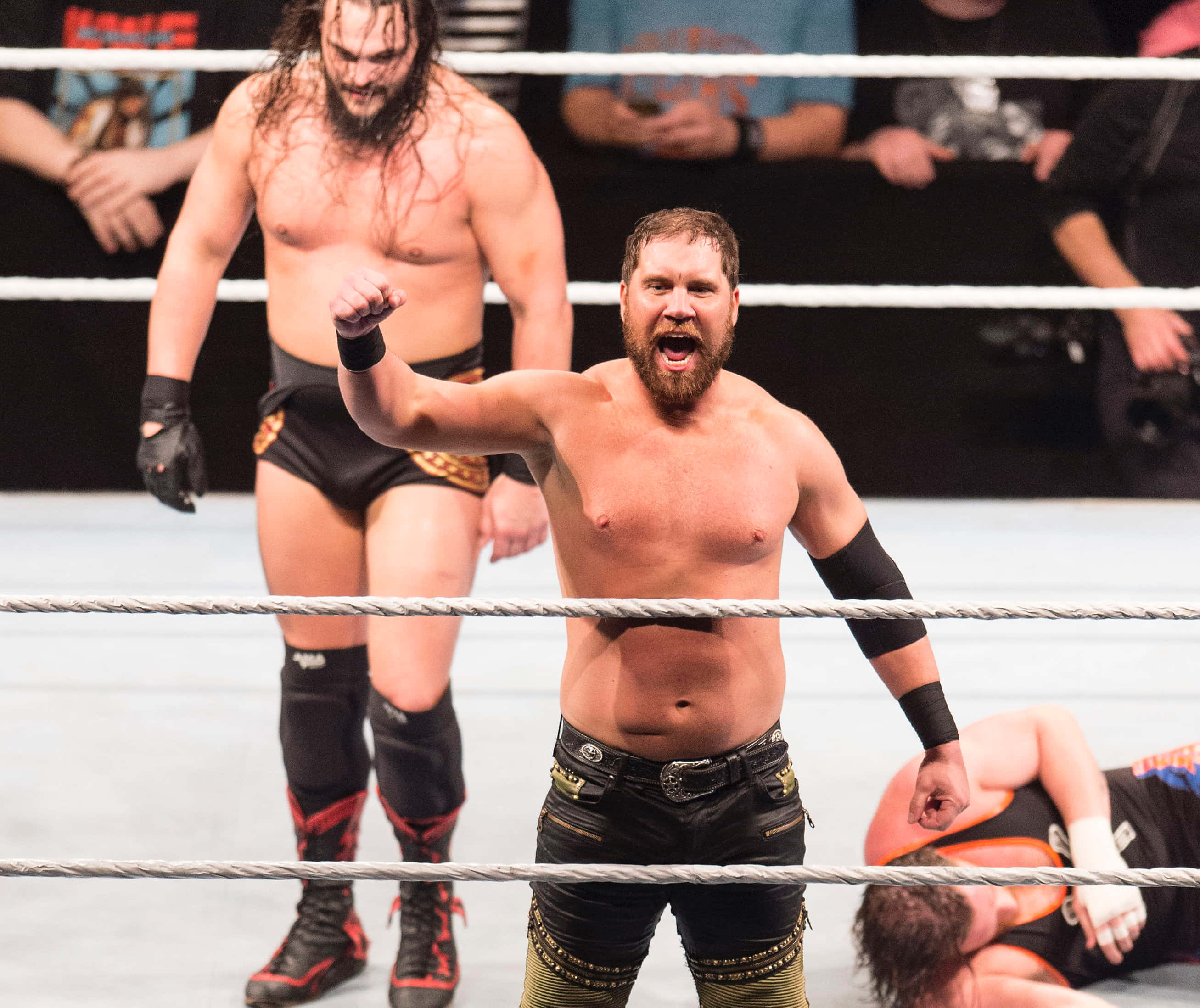 WWE 桌布 壁紙 — 一位留鬍子的摔角手在擂台上高舉拳頭，勝利地咆哮，另一位摔角手站在他身後，第三位則被打敗躺在墊子上。