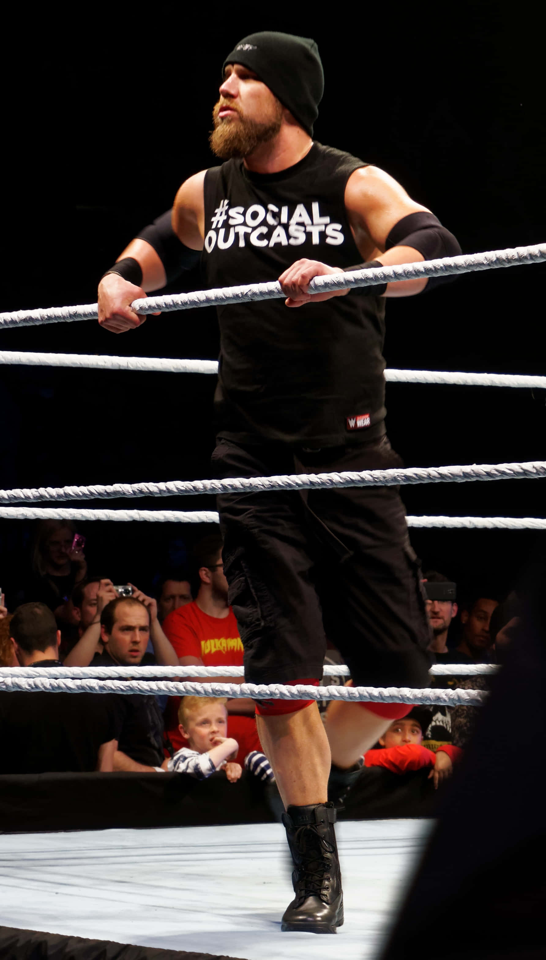 WWE 桌布 壁紙 — 一位留鬍子的摔角手穿著黑色裝備，在明亮的場館燈光下抓住摔角場的繩索，下方有觀眾觀看。