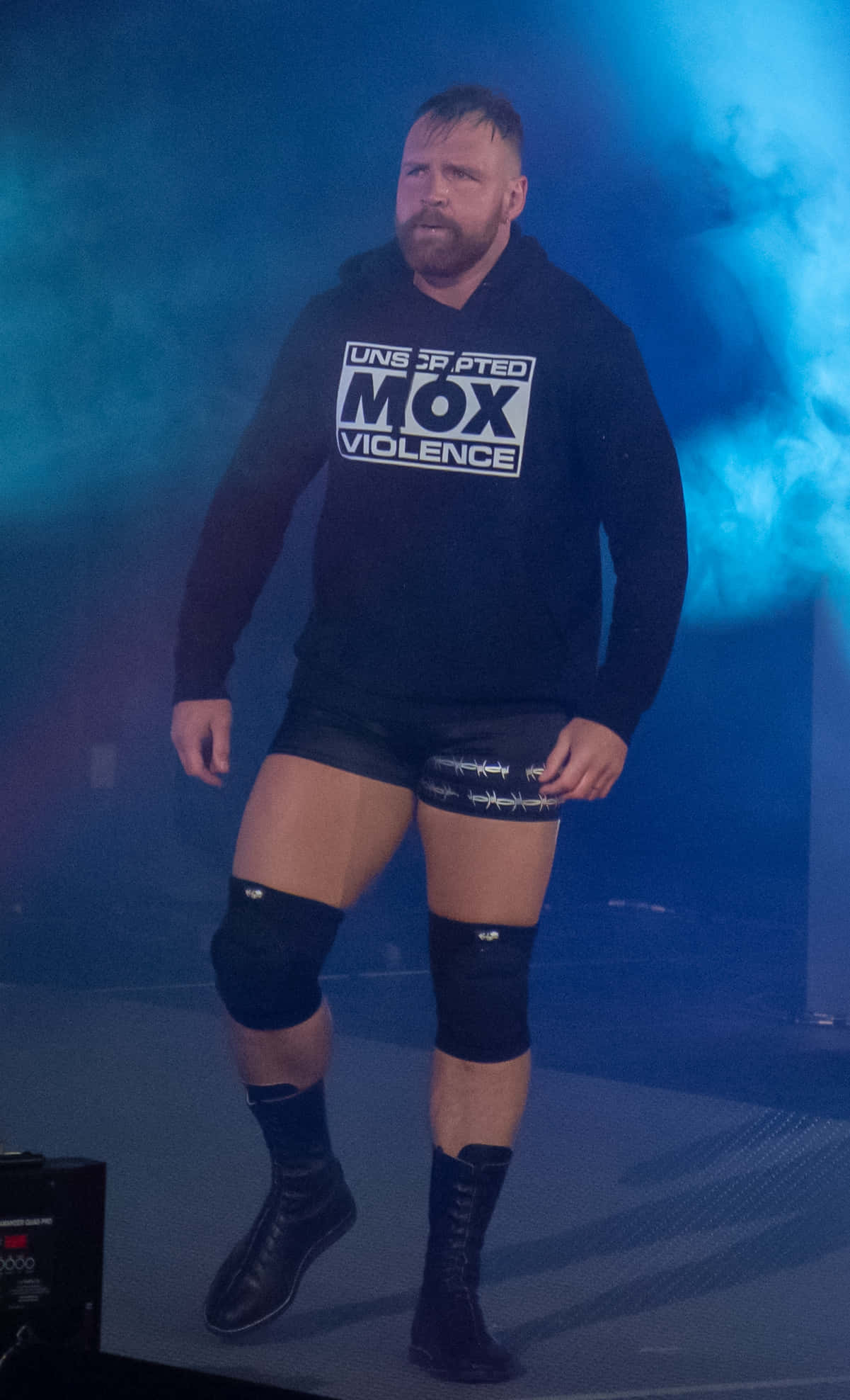 WWE 桌布 壁紙 — 一位留鬍子的摔角手穿著黑色連帽衫和短褲，站在藍色煙霧燈光下的舞台上。