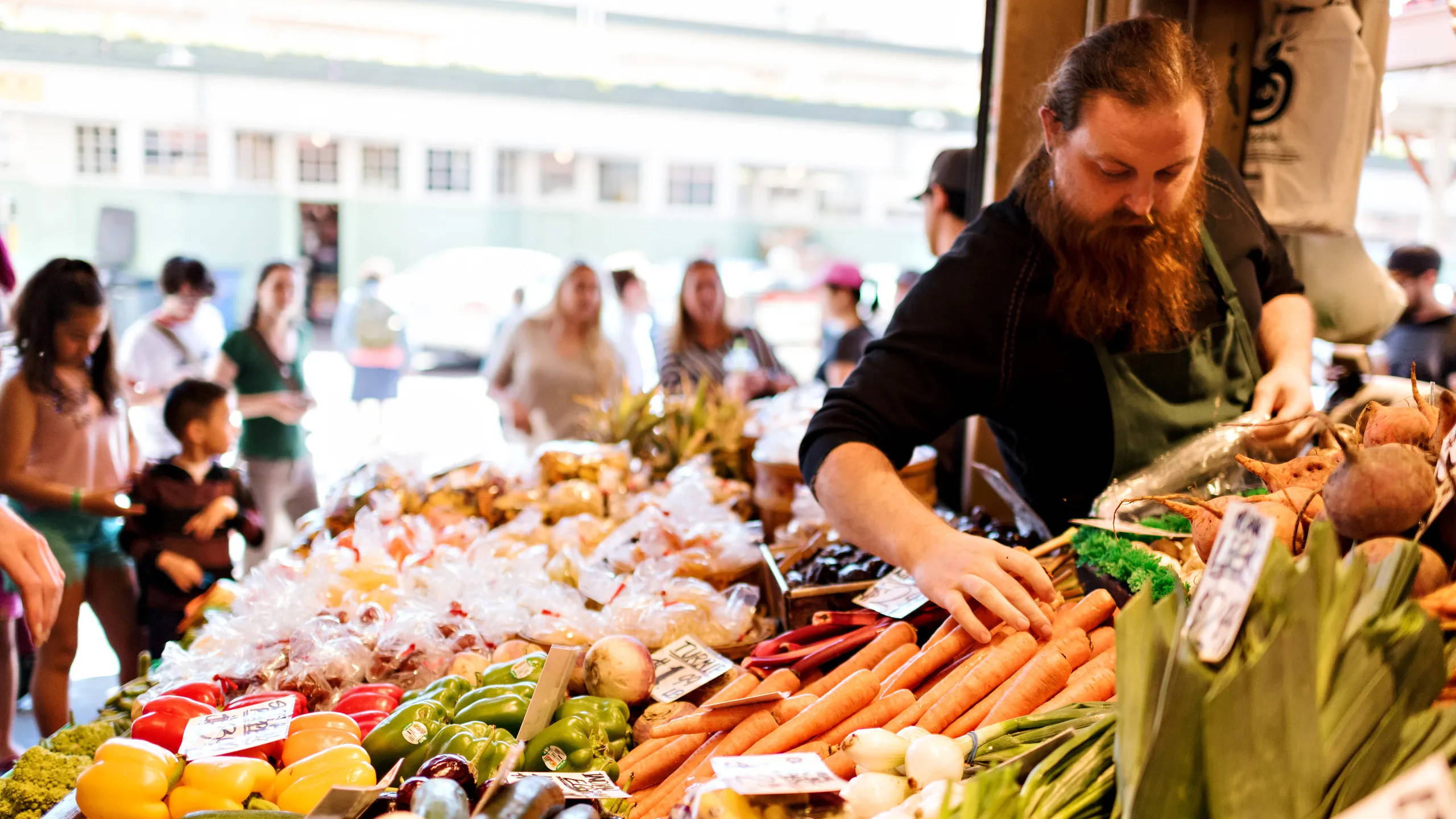 Pike Place Market 桌布 壁紙 — 一位留鬍子的攤販在繁忙的戶外農夫市場攤位上擺放鮮豔的紅蘿蔔和色彩繽紛的農產品。