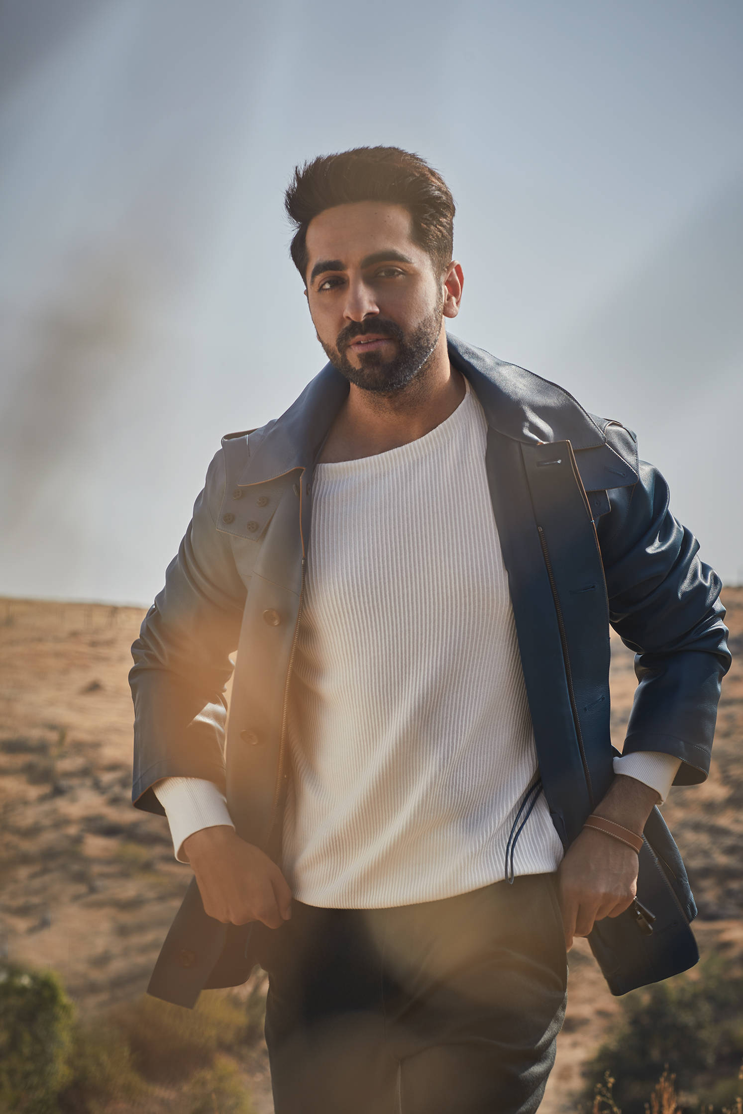 ayushmann khurrana 桌布 壁紙 — 一位留鬍子的時尚男士自信地站在沙漠景觀中，身穿藍色皮夾克和白色羅紋襯衫。