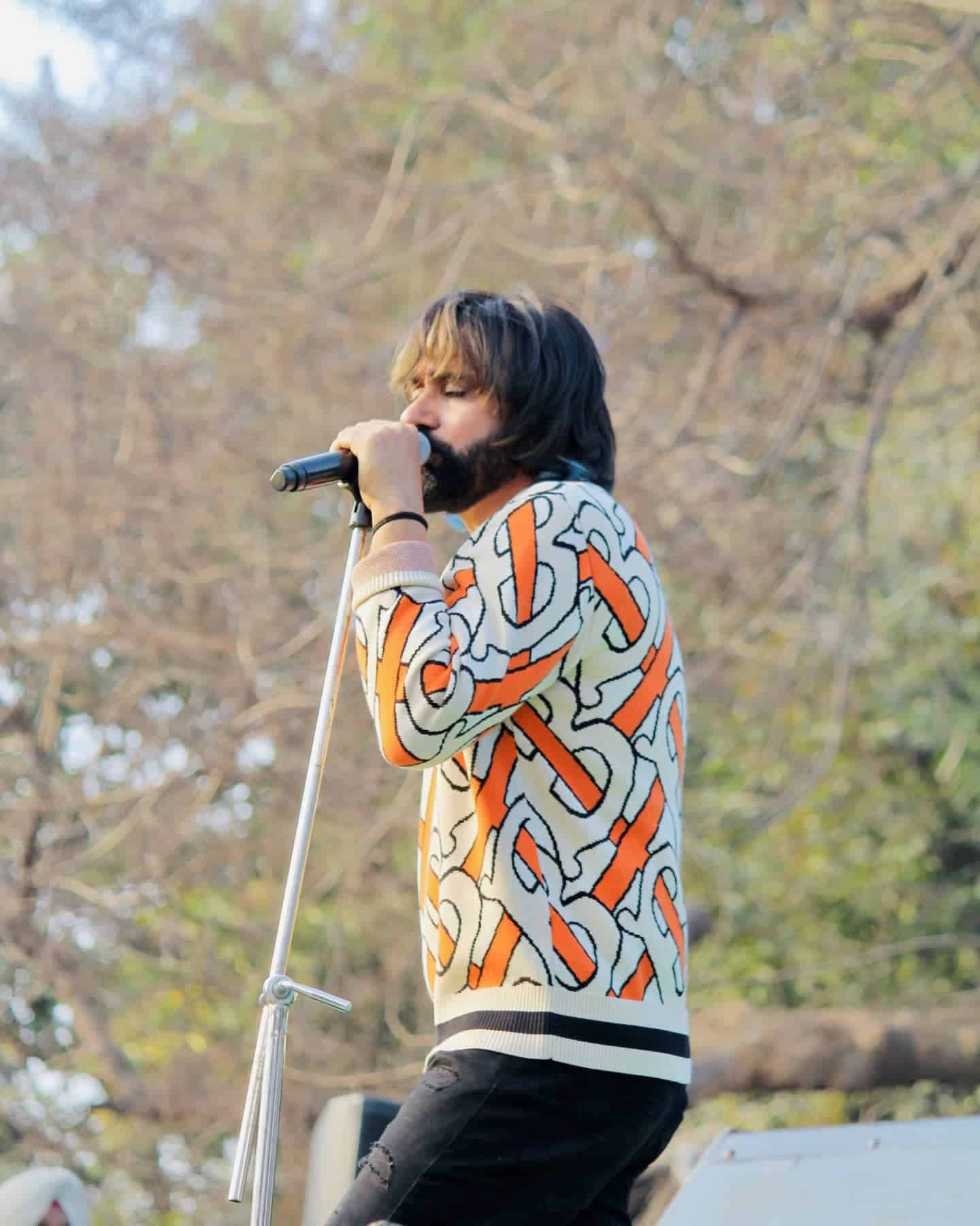 Singing 桌布 壁紙 — 一位留鬍子的歌手穿著大膽的橙色和白色圖案毛衣，在戶外熱情地對著麥克風唱歌。